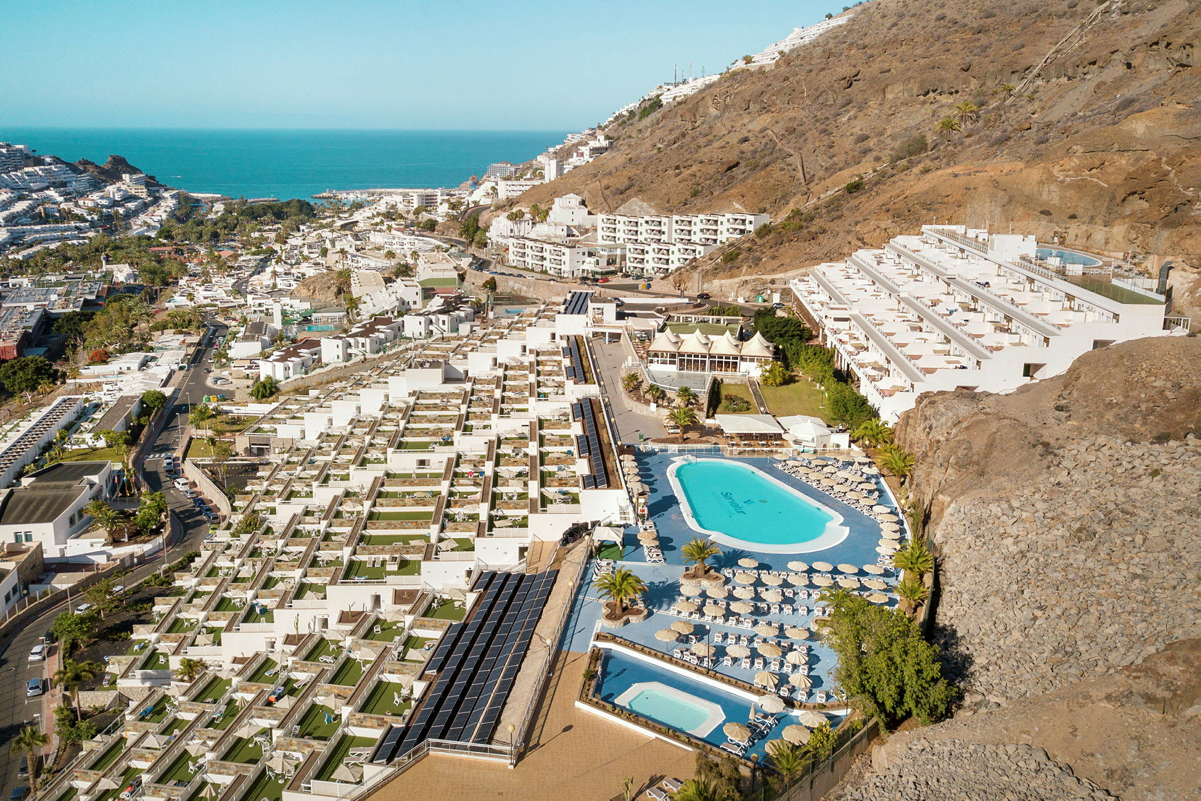 Flygfoto av en hotellanläggning med pooler, solstolar och terrasser, belägen vid en sluttning nära kusten. (AI-genererad text)
