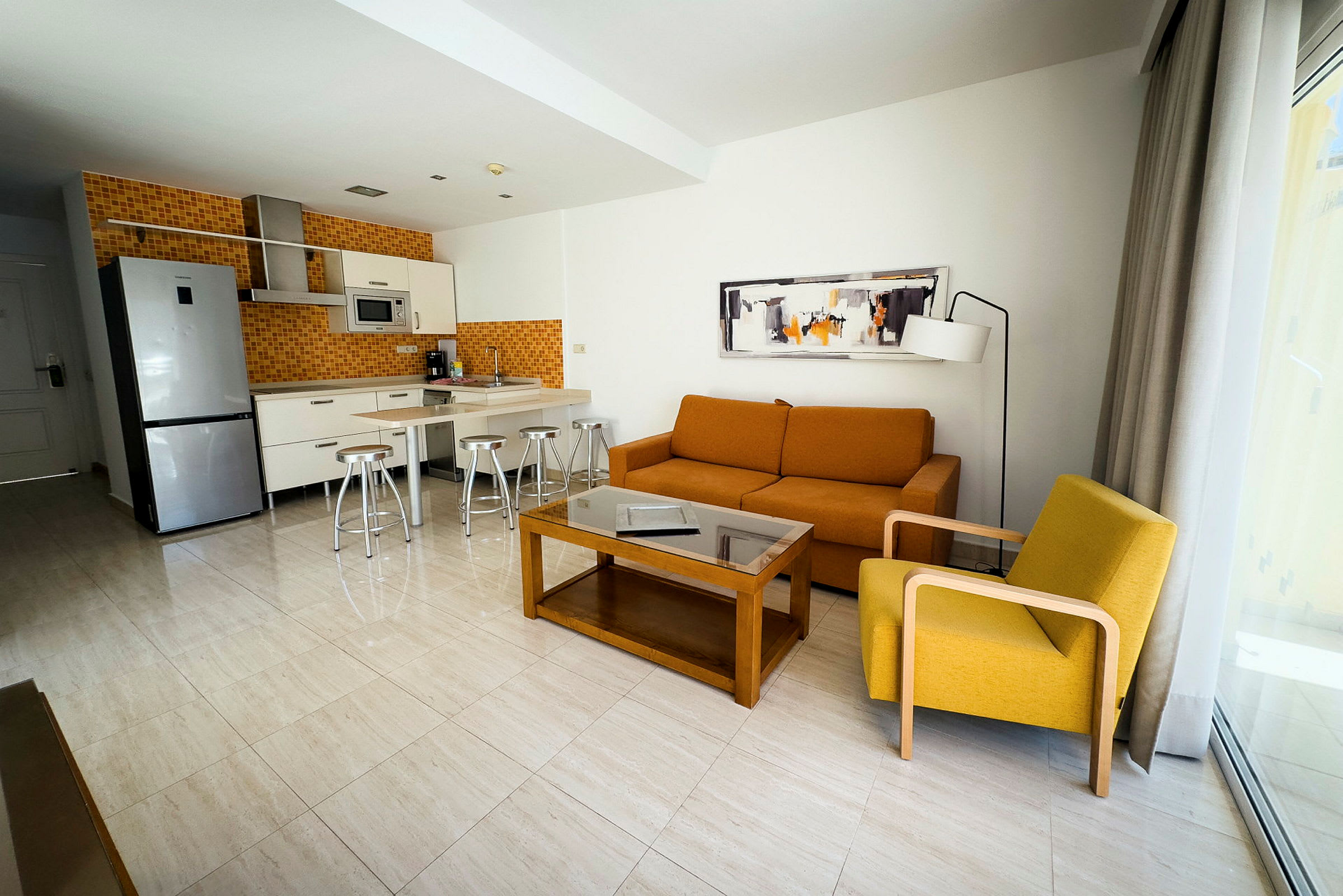 Modernt hotellrum med kök och vardagsrumsmöbler inklusive orange soffa och gul fåtölj. (AI-genererad text)
