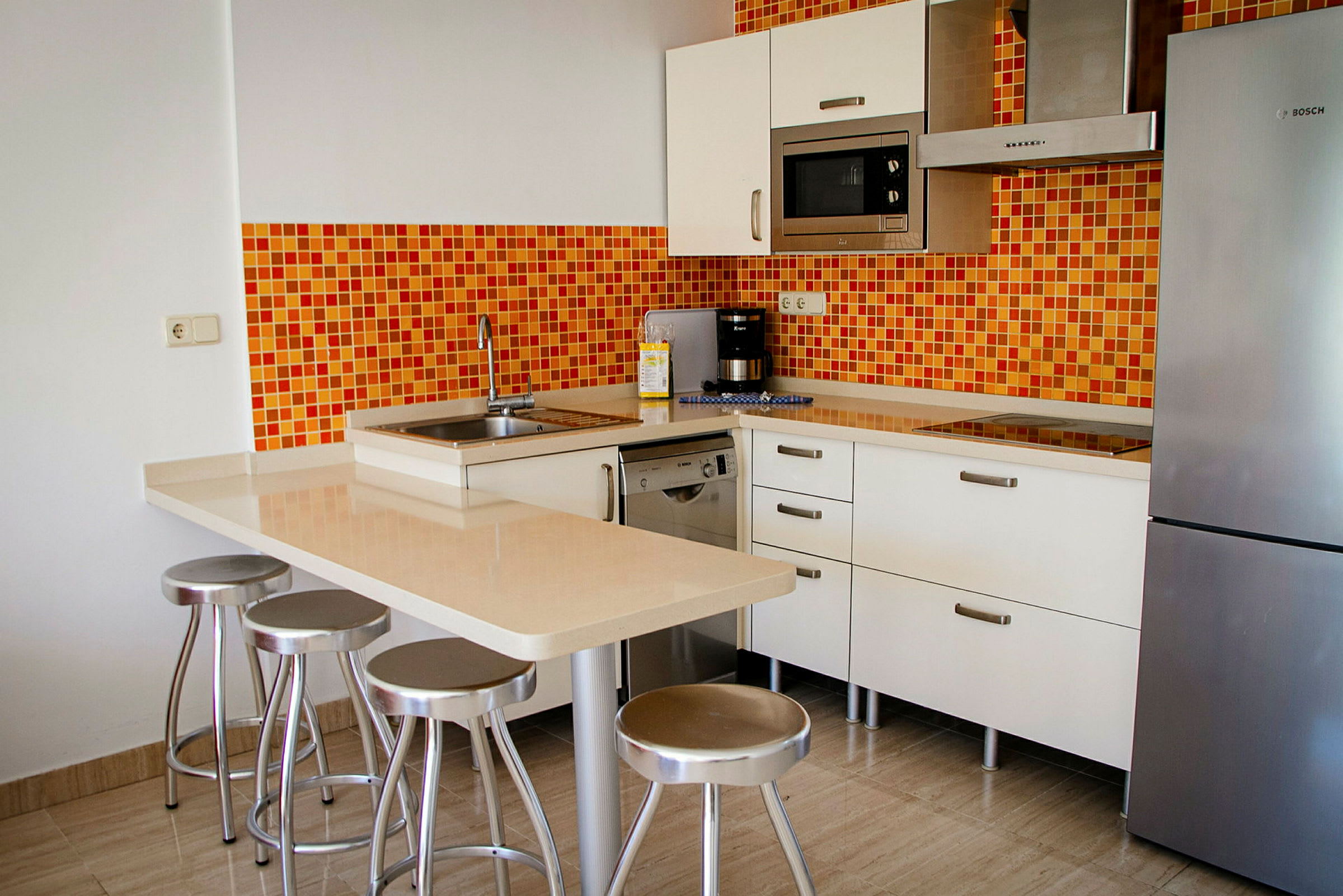 Modernt kök med orange mosaikvägg, kylskåp, mikrovågsugn och köksön med barstolar. (AI-genererad text)