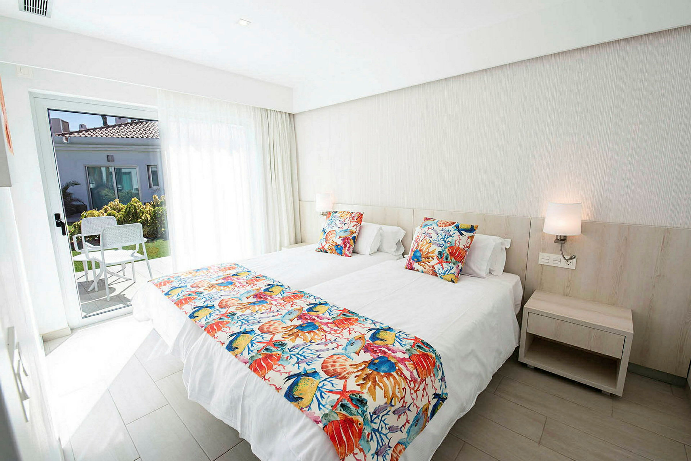 Ett hotellrum med två enkelsängar, färgglada kuddlakan och en dörr till en terrass. (AI-genererad text)