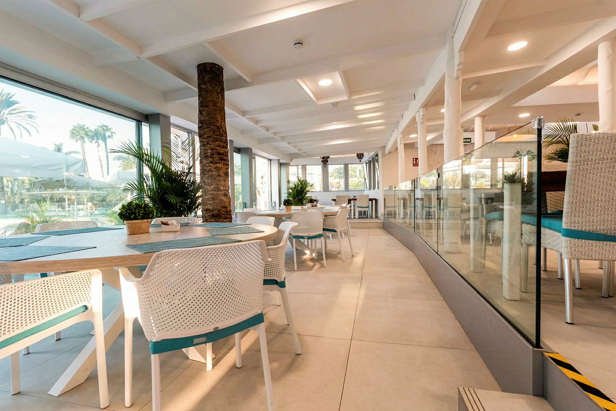 Interiören av en restaurang med vita stolar och bord, glasväggar och utsikt mot grönska utomhus. (AI-genererad text)