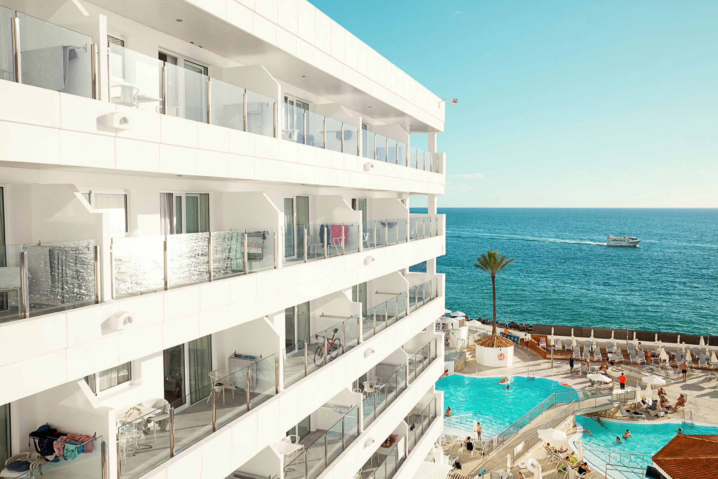 Utsikt över ett modernt hotell med balkonger, en utomhuspool och havet i bakgrunden. (AI-genererad text)