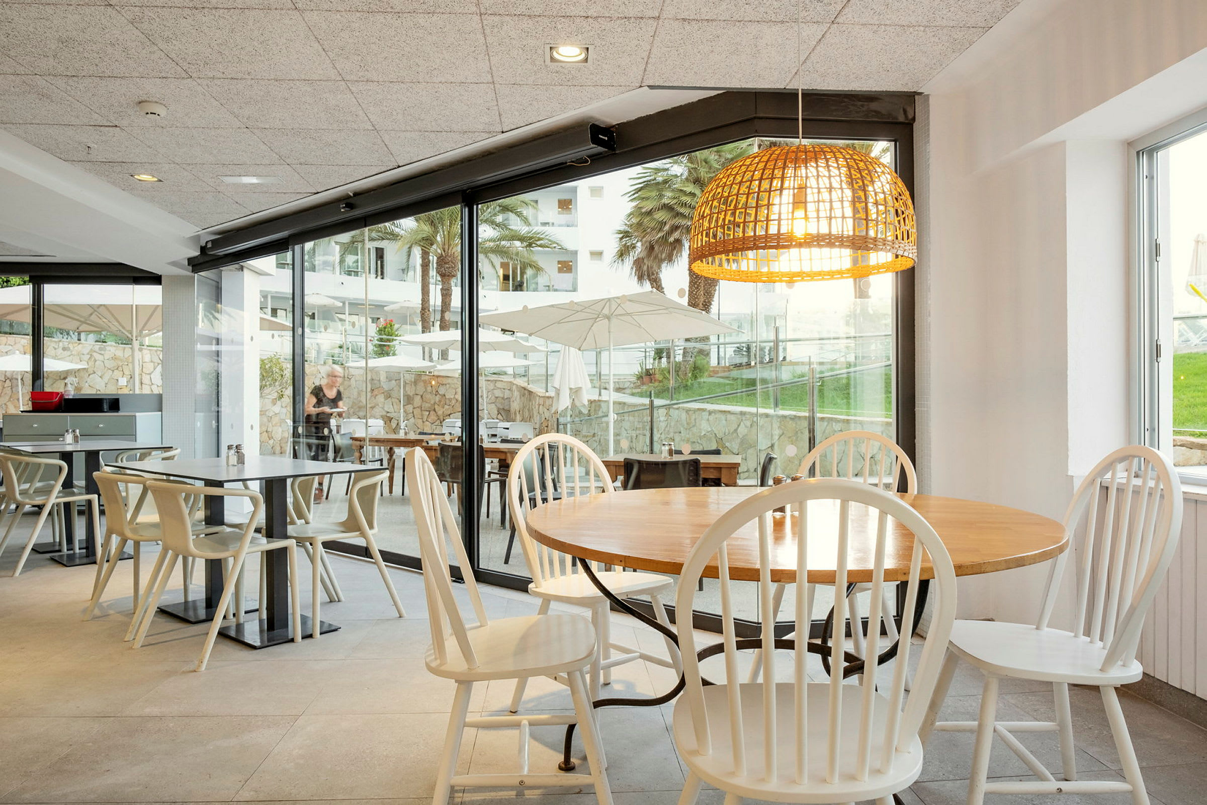 En restaurang med bord och stolar. Stora glasfönster visar utsikten med parasoller, grönområden och palmer. (AI-genererad text)