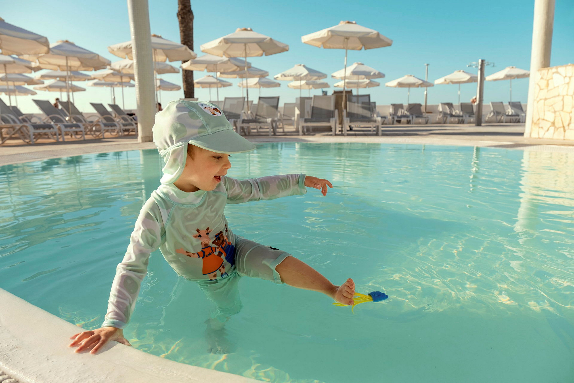 Ett barn leker i en grund pool med flera solstolar och parasoller i bakgrunden. (AI-genererad text)