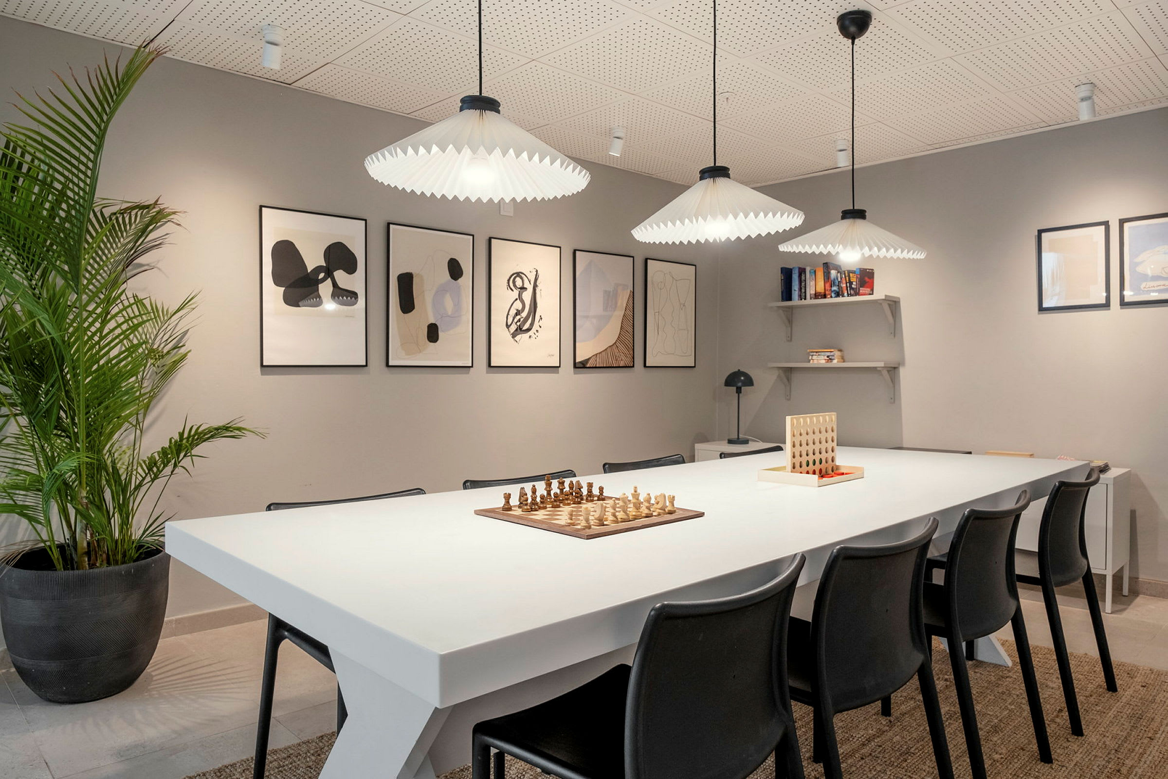 Bild av ett rum med en vit bord omgiven av svarta stolar. På bordet står schack och ett spel med brickor. Väggarna dekoreras med inramade abstrakta konstverk, och en hylla innehåller böcker. (AI-genererad text)