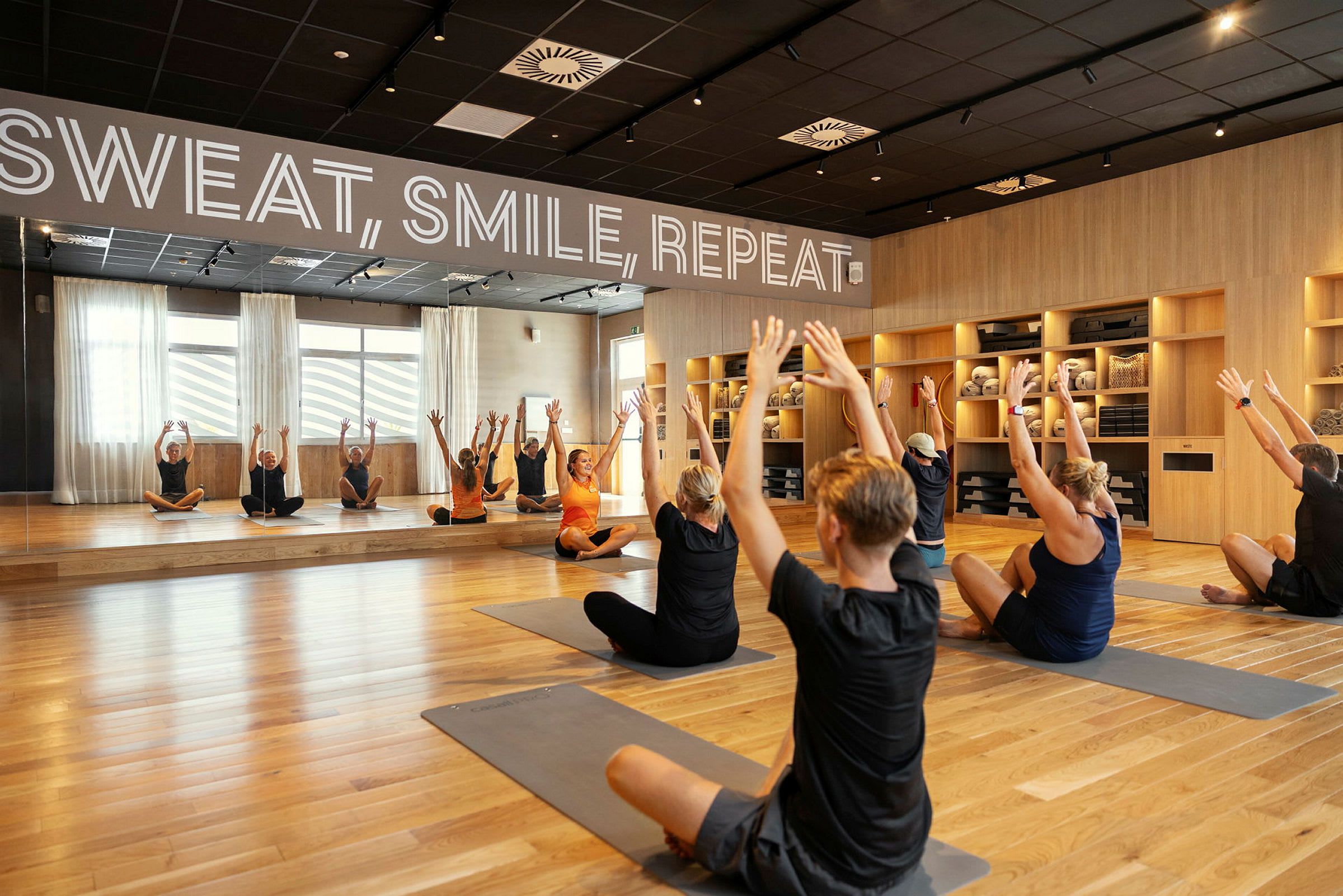 En träningssal med spegelvägg där flera personer utför stretchingövningar på yogamattor. (AI-genererad text)