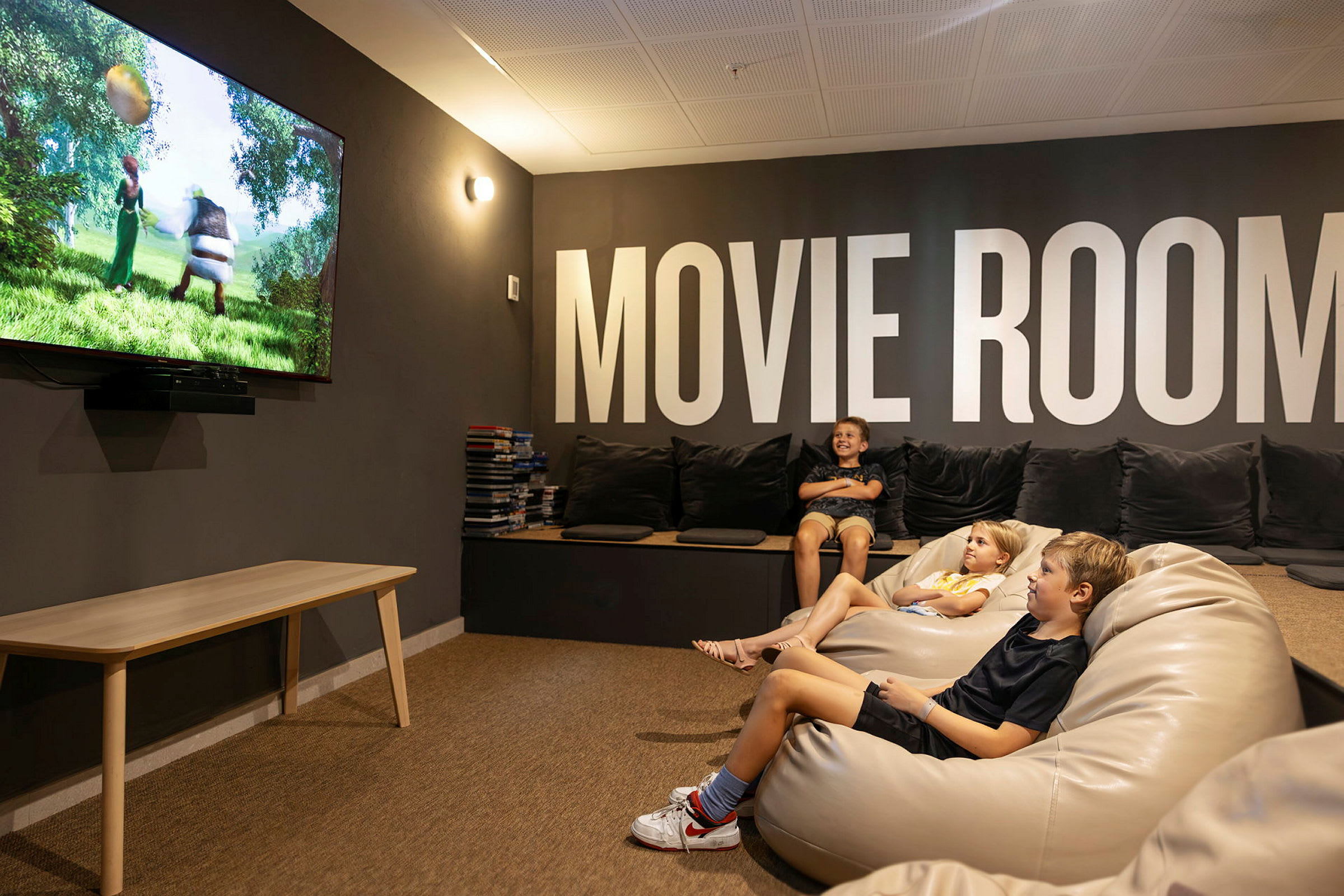 Ett rum med mjuka sittmöbler och en stor TV som visar en animerad film. Väggarna pryds av texten "MOVIE ROOM". (AI-genererad text)