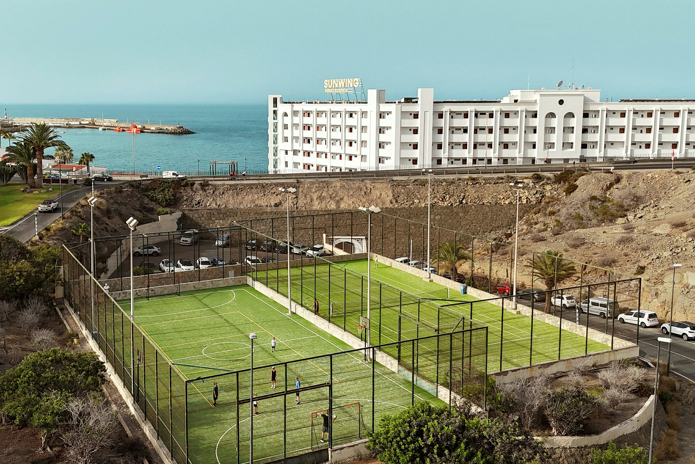 Fotbollsplan med grönt konstgräs vid kusten, byggnad i bakgrunden. (AI-genererad text)