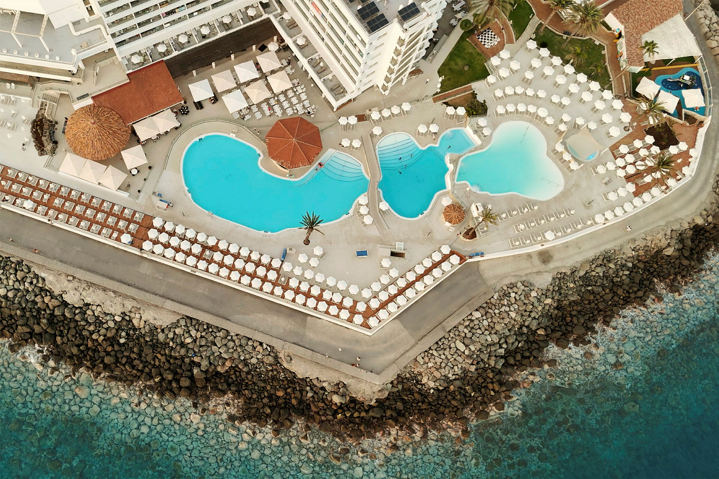 Flygfoto över ett hotellområde med flera pooler nära havet. (AI-genererad text)