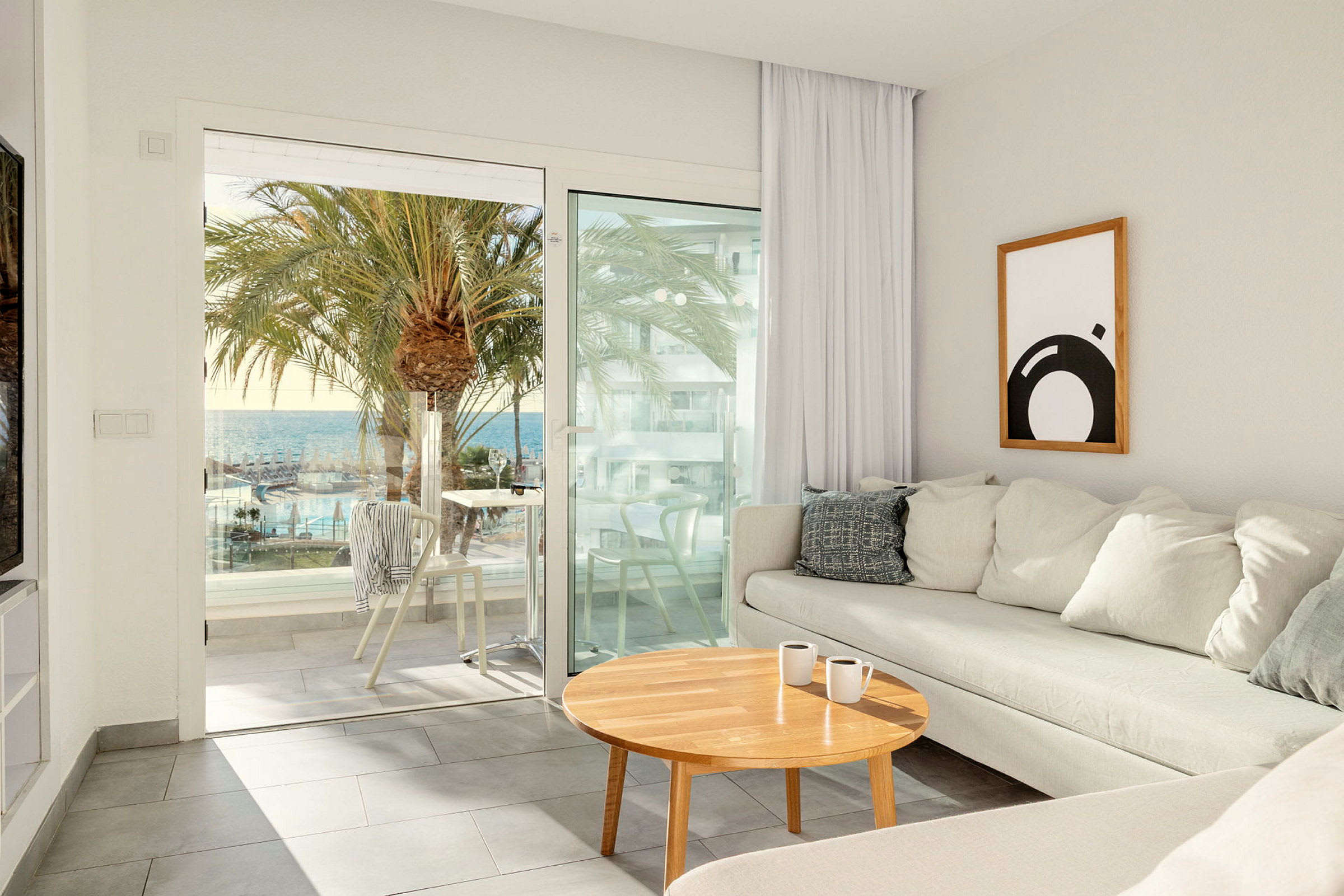 Ett ljust vardagsrum, med en beige soffa och ett träbord, har utsikt genom glasdörrar mot en terrass med utsikt över palmer, en pool och havet. (AI-genererad text)
