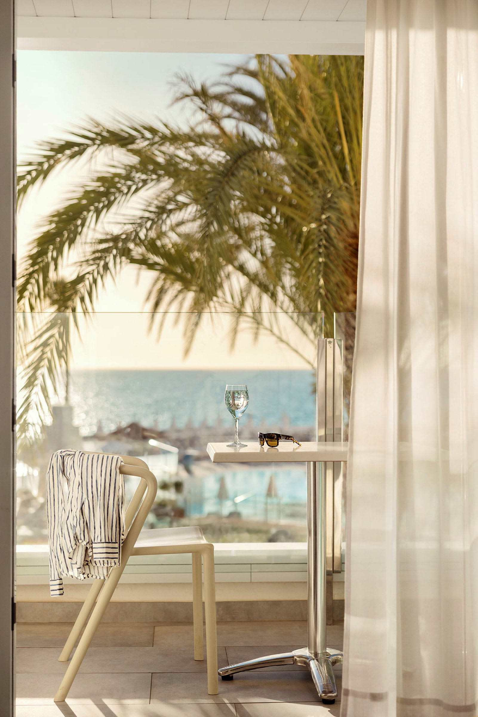Ett bord med ett glas vin och ett par solglasögon står på en terrass med utsikt över stranden och havet. En stol har en randig skjorta hängande över ryggen och står bredvid bordet. (AI-genererad text)