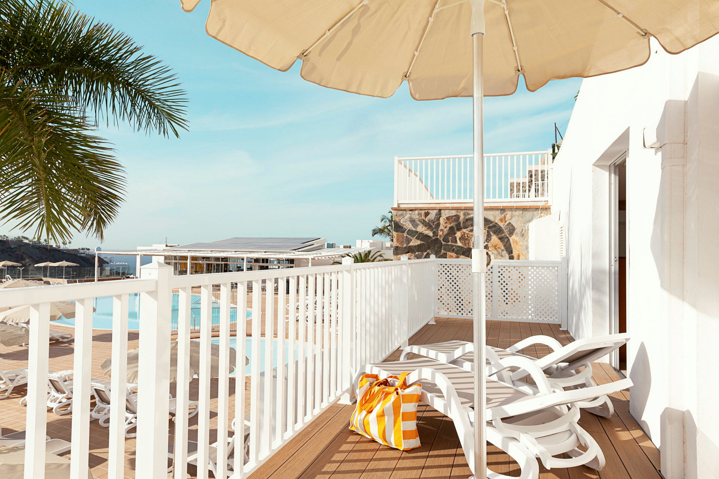 Vy över en terrass med solstolar, parasoll och en strandväska. I bakgrunden syns en pool och palmer. (AI-genererad text)