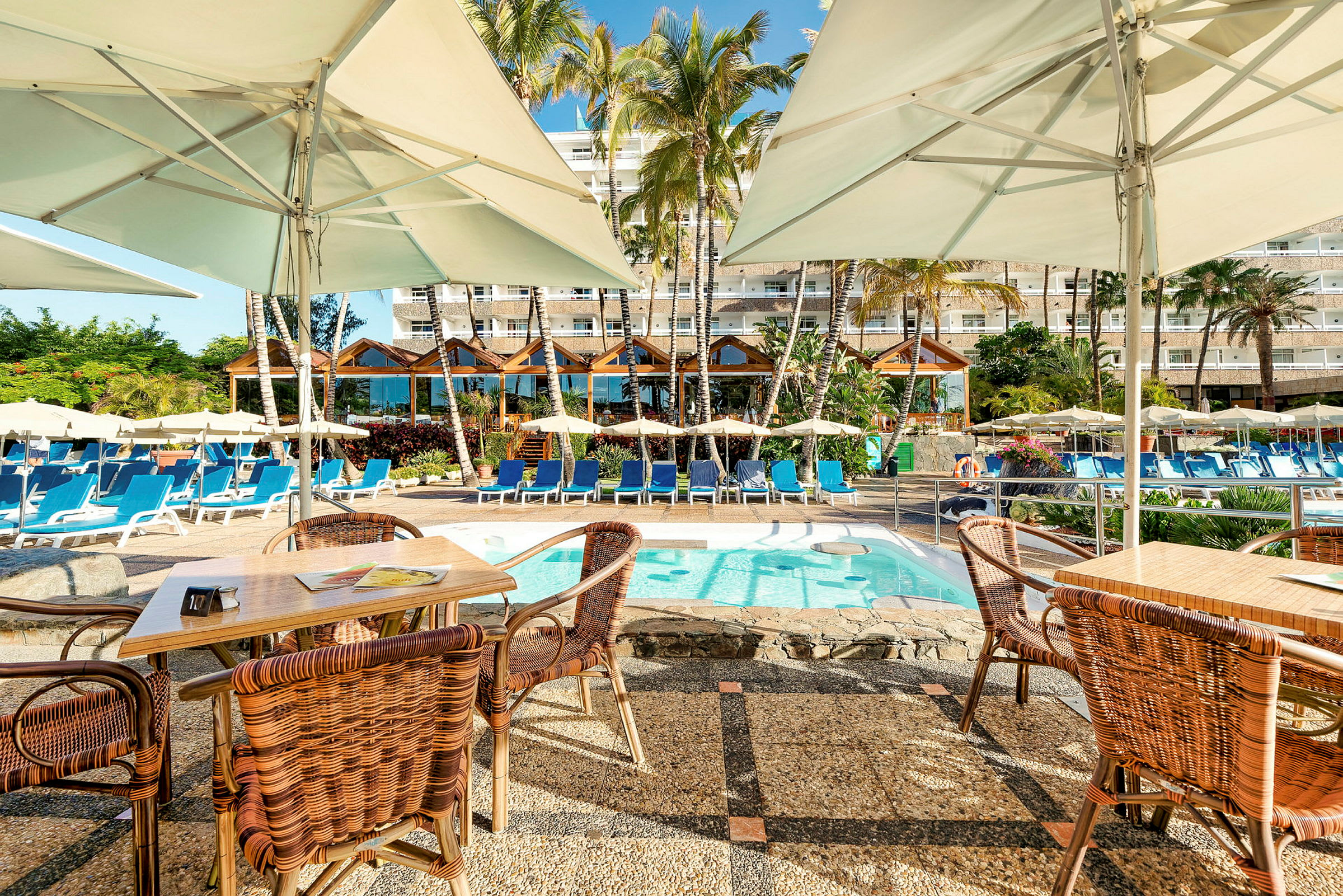 En pool med solstolar och parasoller, omgivet av palmer och ett hotell i bakgrunden. (AI-genererad text)