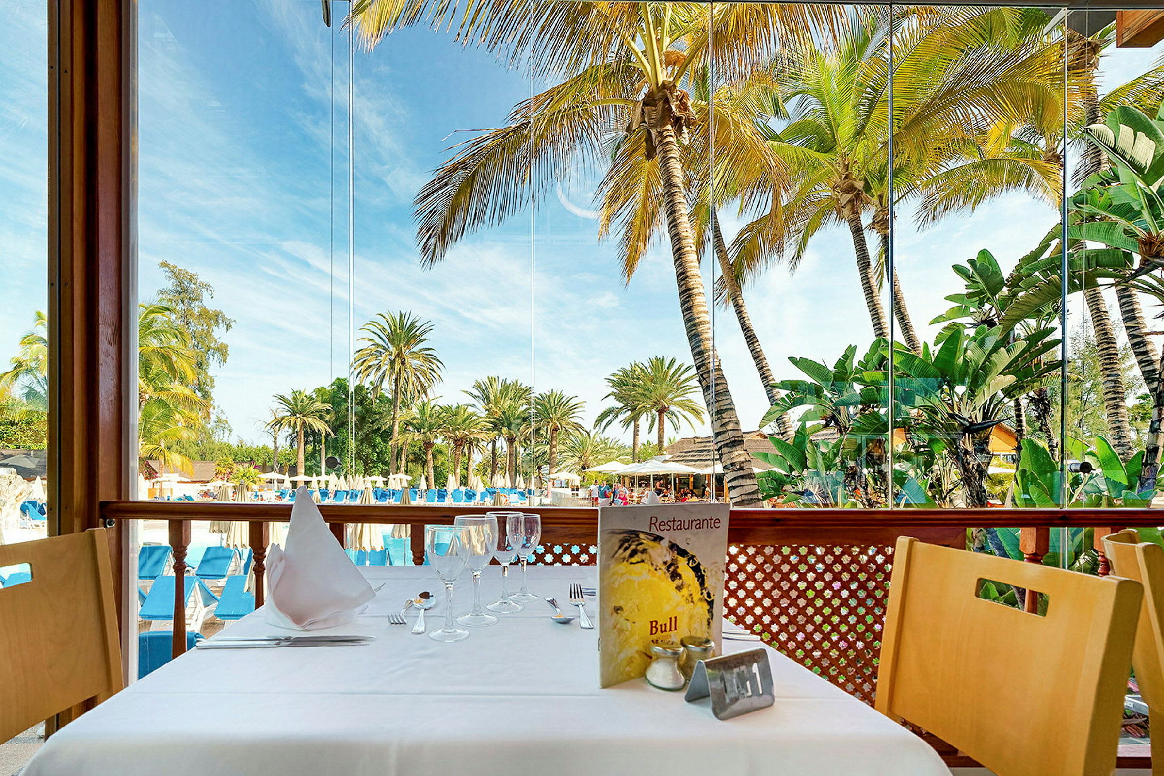 En dukad restaurangbord med utsikt över palmträd och en pool. (AI-genererad text)