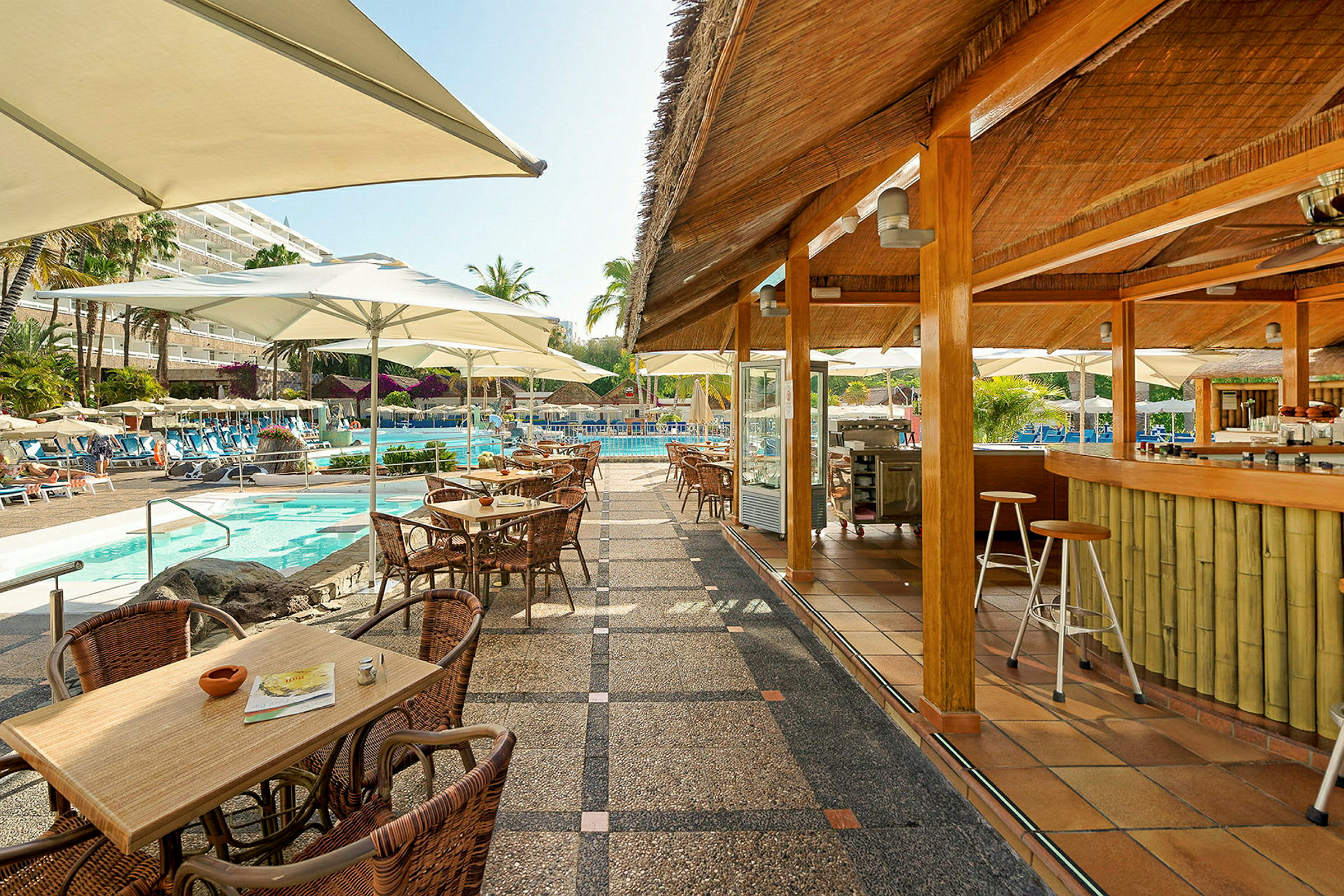 En poolbar med uteservering och bord, stolar samt parasoller vid en poolområde med solstolar och växter. (AI-genererad text)