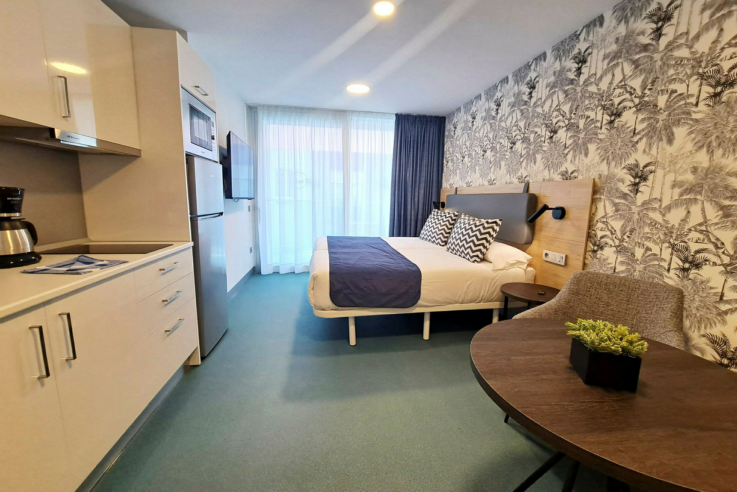 Ett hotellrum med dubbelsäng, ett kökshörn och en sittplats. Väggarna har mönstrad tapet. (AI-genererad text)