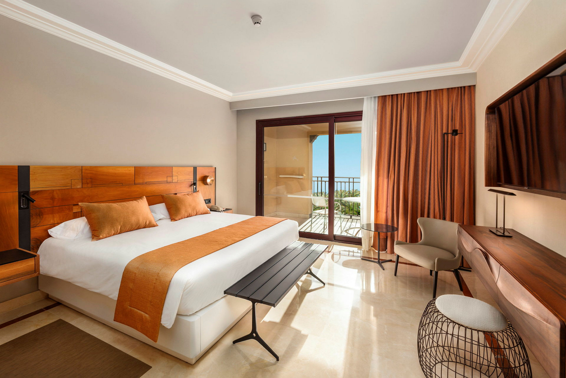 Ett hotellrum med en stor säng, orange inredningsdetaljer, ett skrivbord och en balkong med havsutsikt. (AI-genererad text)