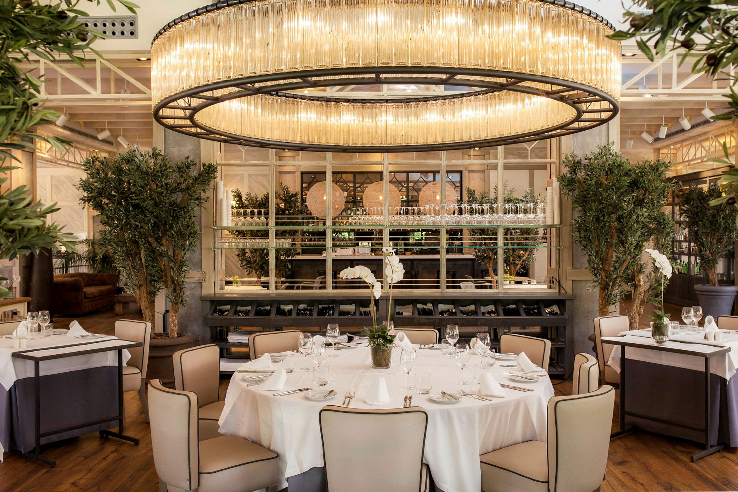 Elegant restauranginteriör med en rund lampa i taket ovanför ett dukat bord omgivet av stolar. Rummet har också stora växter och en modern dekor. (AI-genererad text)