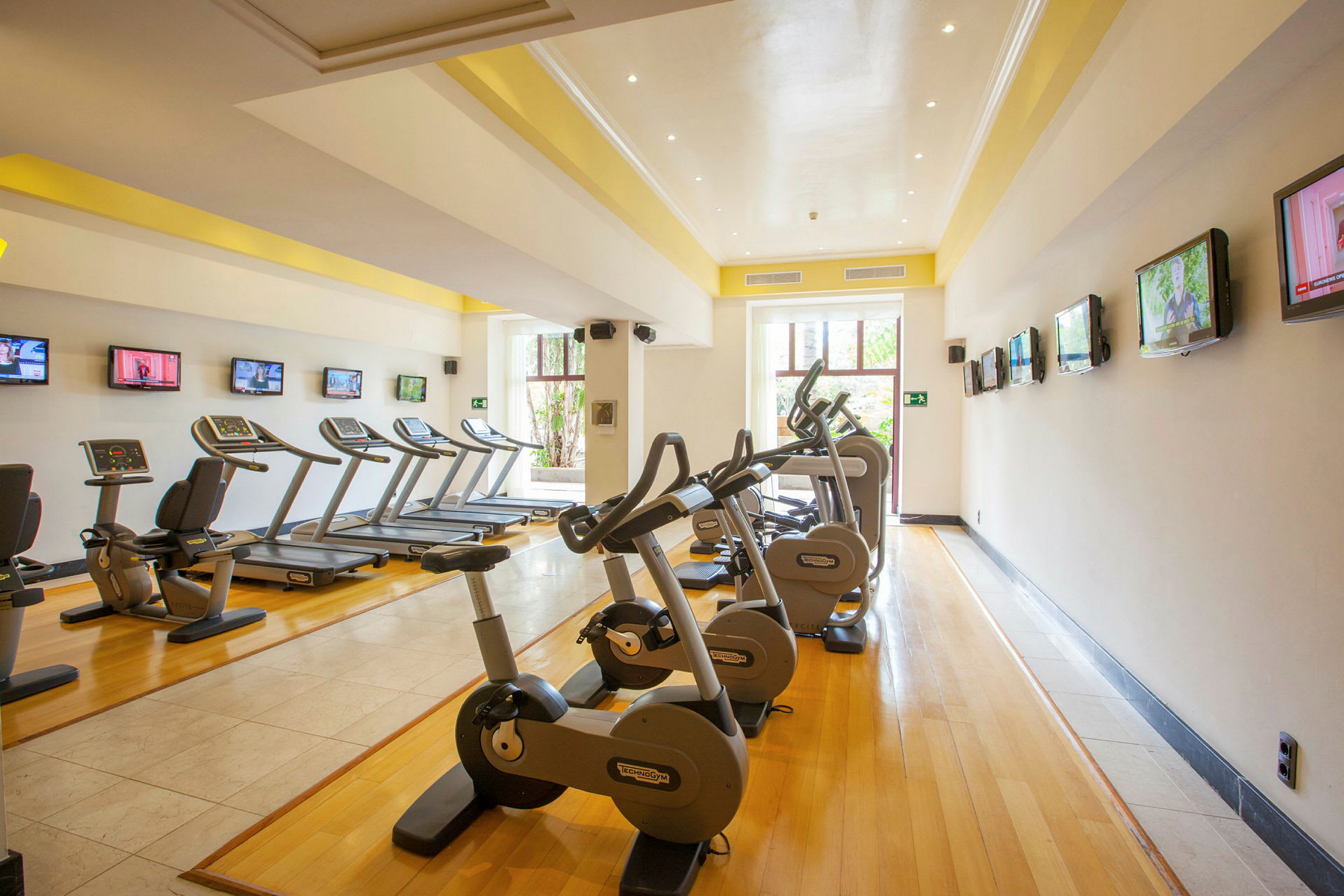 Bild på ett gym som visar motionscyklar och löpband uppradade längs ett rum med trägolv och flera TV-skärmar på väggarna. (AI-genererad text)