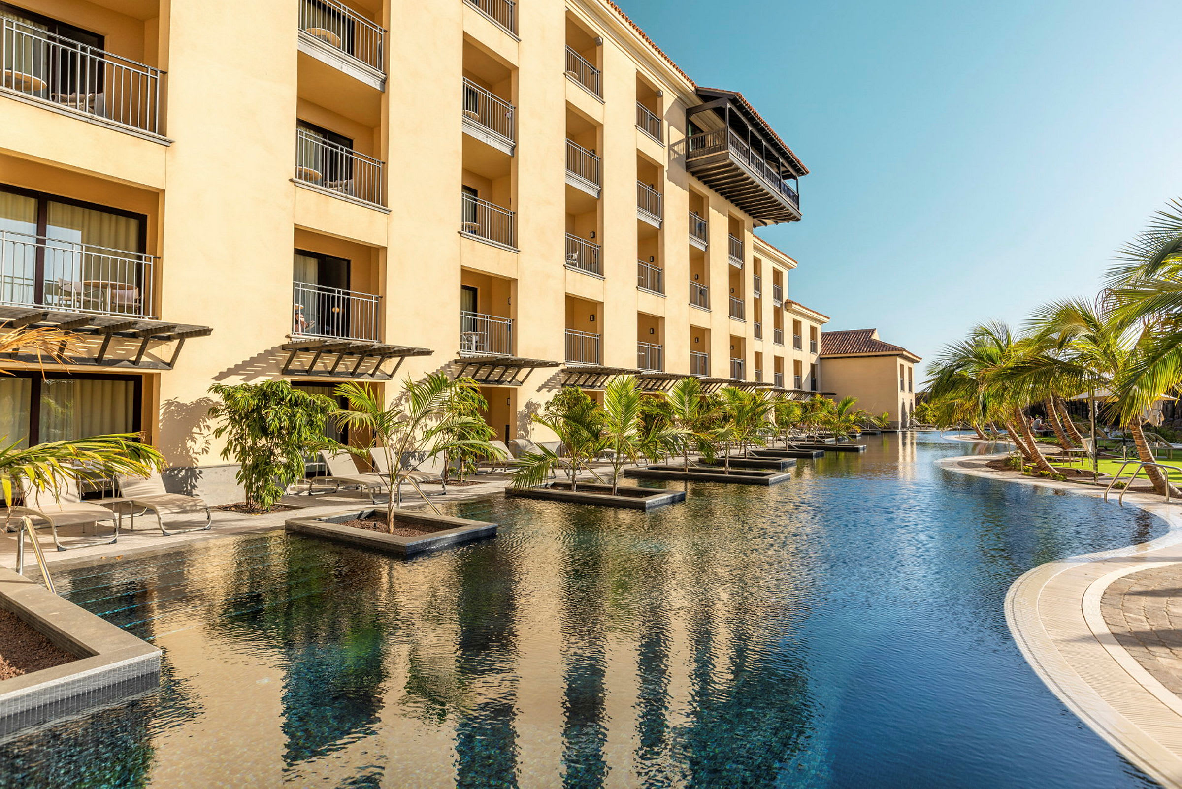 Ett hotellkomplex med en pool omgiven av solstolar och palmer. (AI-genererad text)