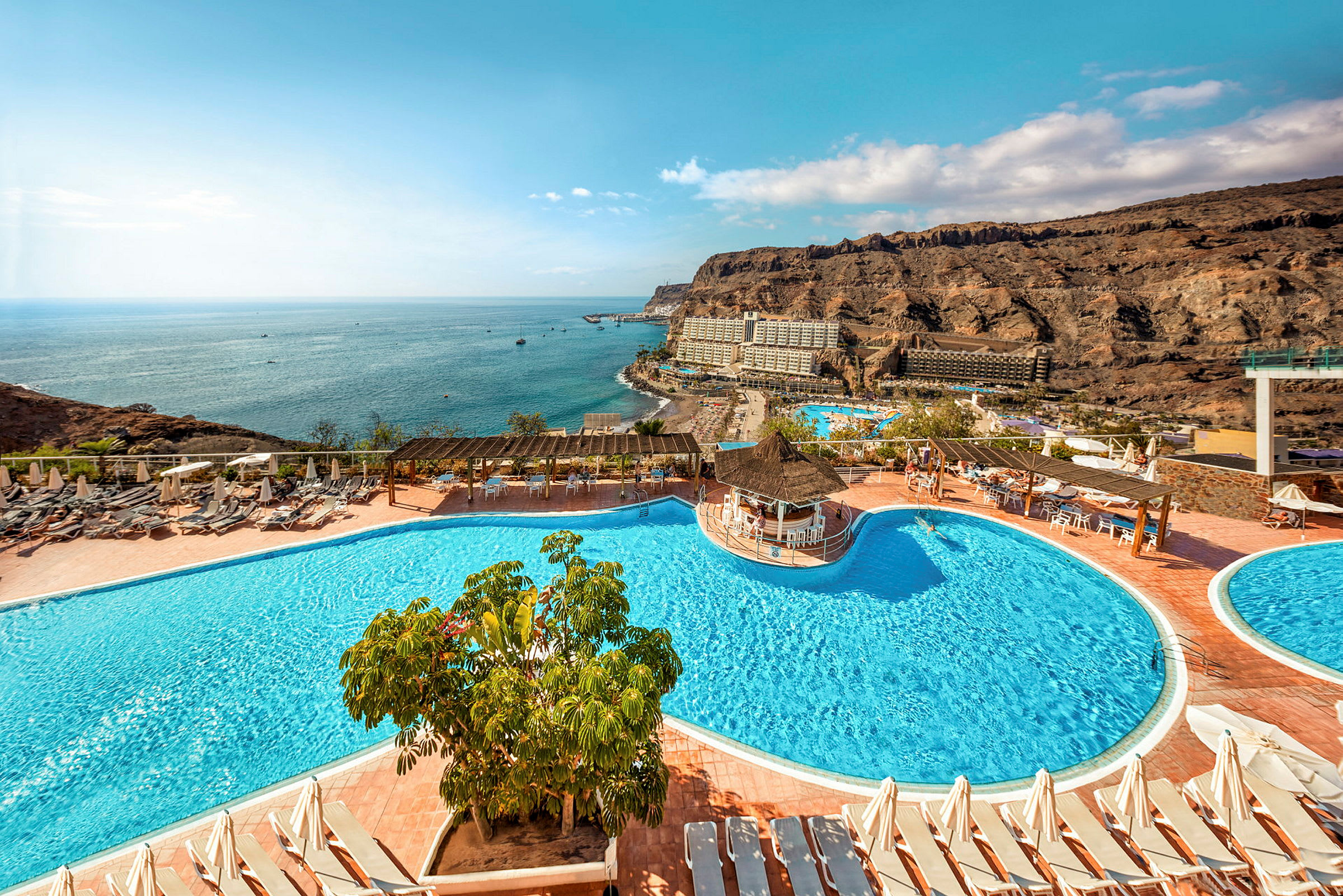 Hotellområde med pooler, solstolar och utsikt över havet samt omgivande klippor. (AI-genererad text)
