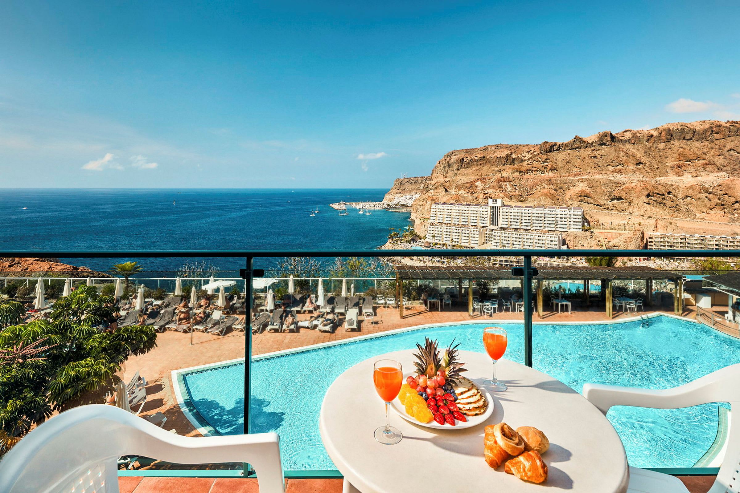 Utsikt över en pool, tallrik med frukt och dryck på en terrass med hav och berg i bakgrunden. (AI-genererad text)