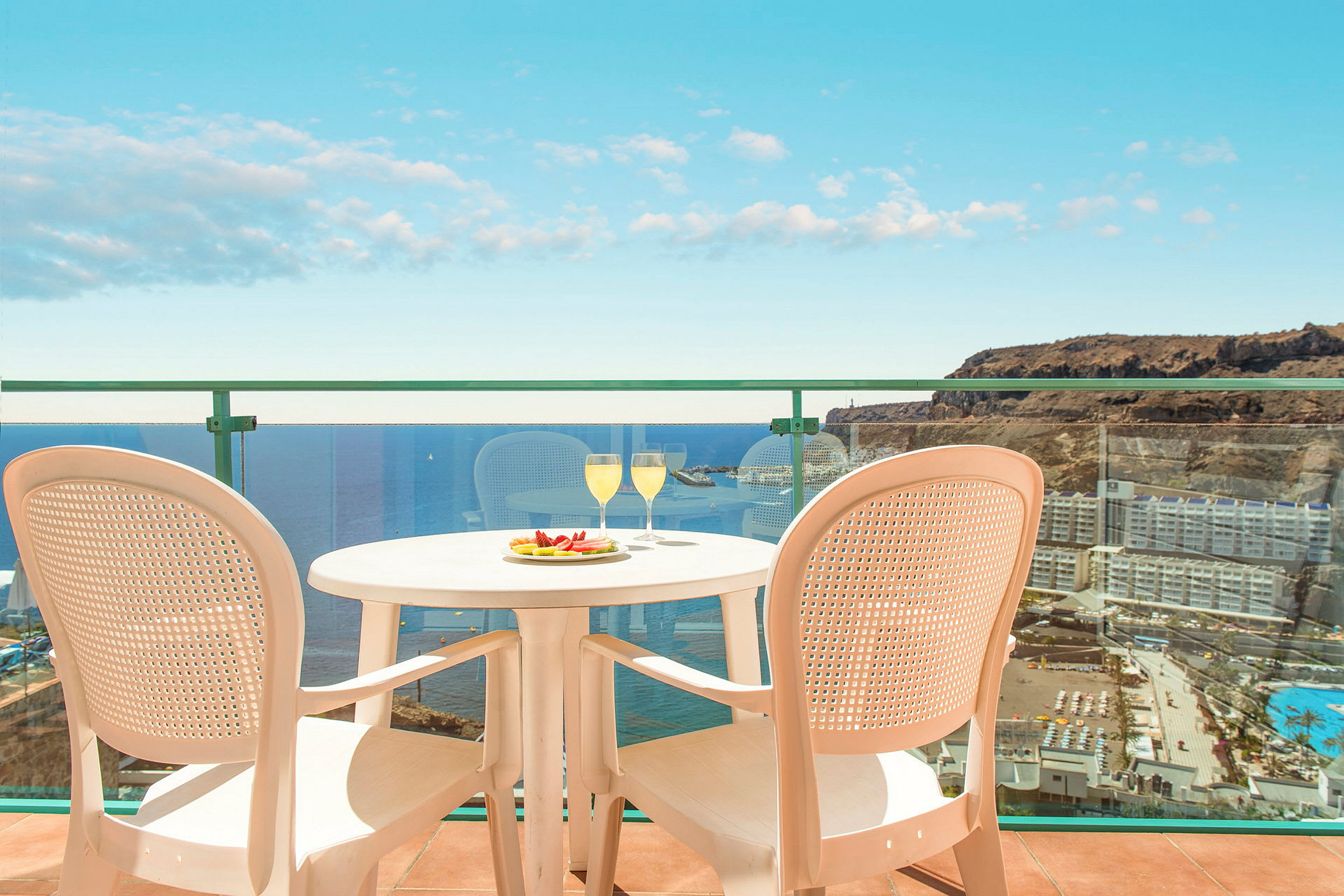 Bild av en balkong med utsikt över havet och omgivande byggnader. På balkongen står ett bord med två glas och en tallrik med frukt samt två stolar. (AI-genererad text)
