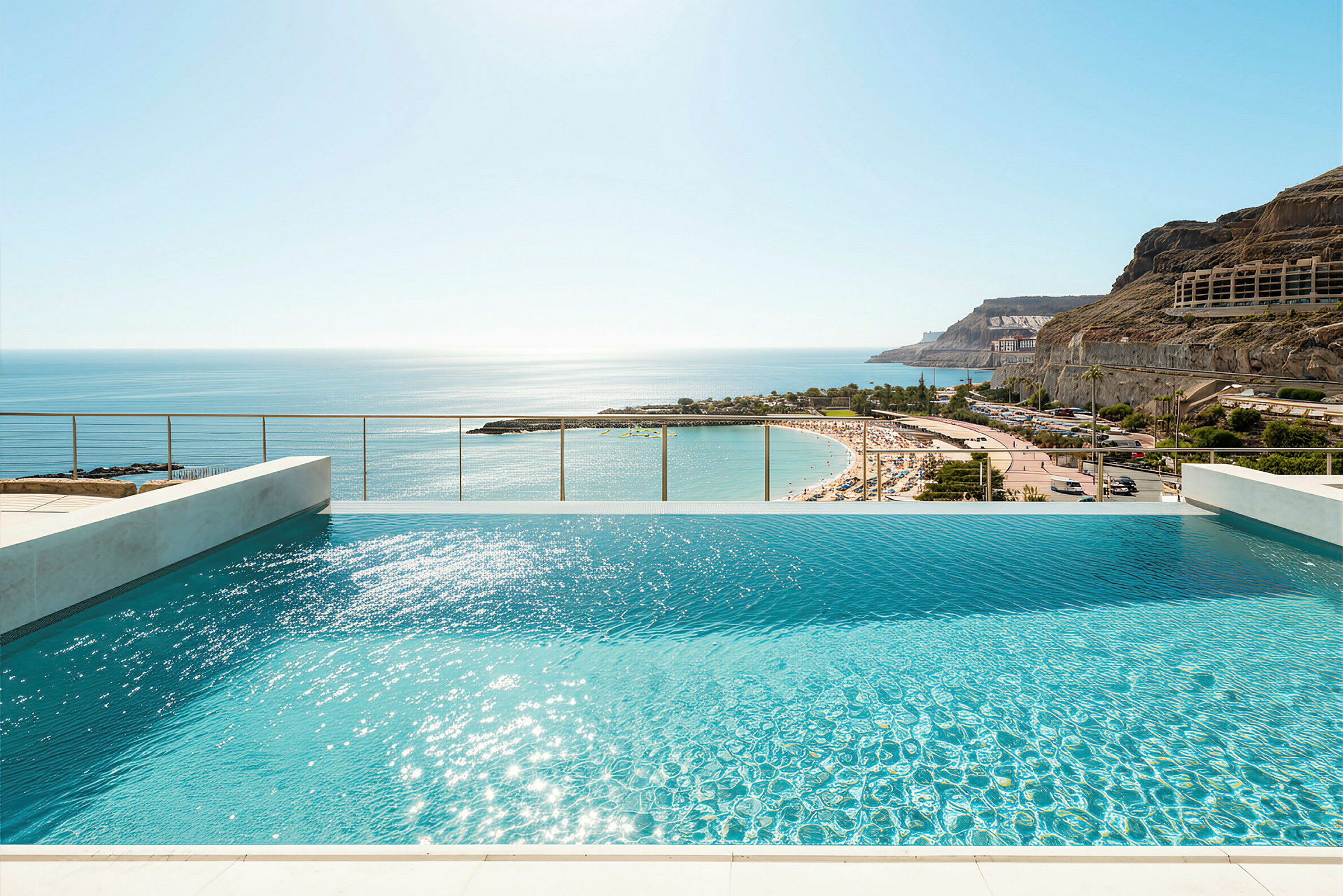 Utsikt över en infinitypool med solblänk på vattenytan och en sandstrand i bakgrunden. (AI-genererad text)