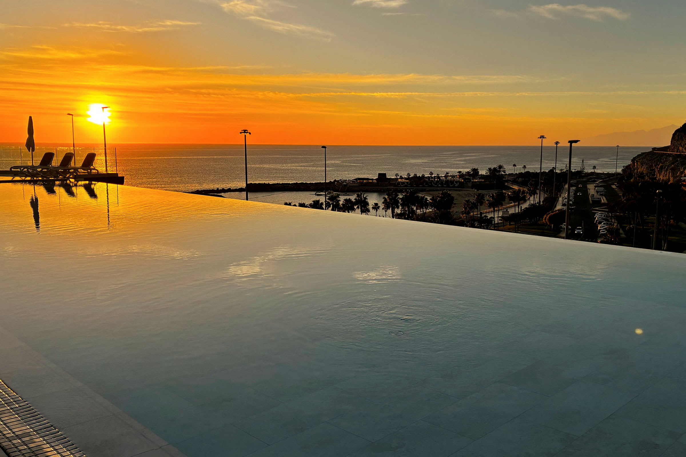 En infinitypool med utsikt över havet vid solnedgången. Solen speglas i vattnet och palmer skymtar i fjärran. (AI-genererad text)