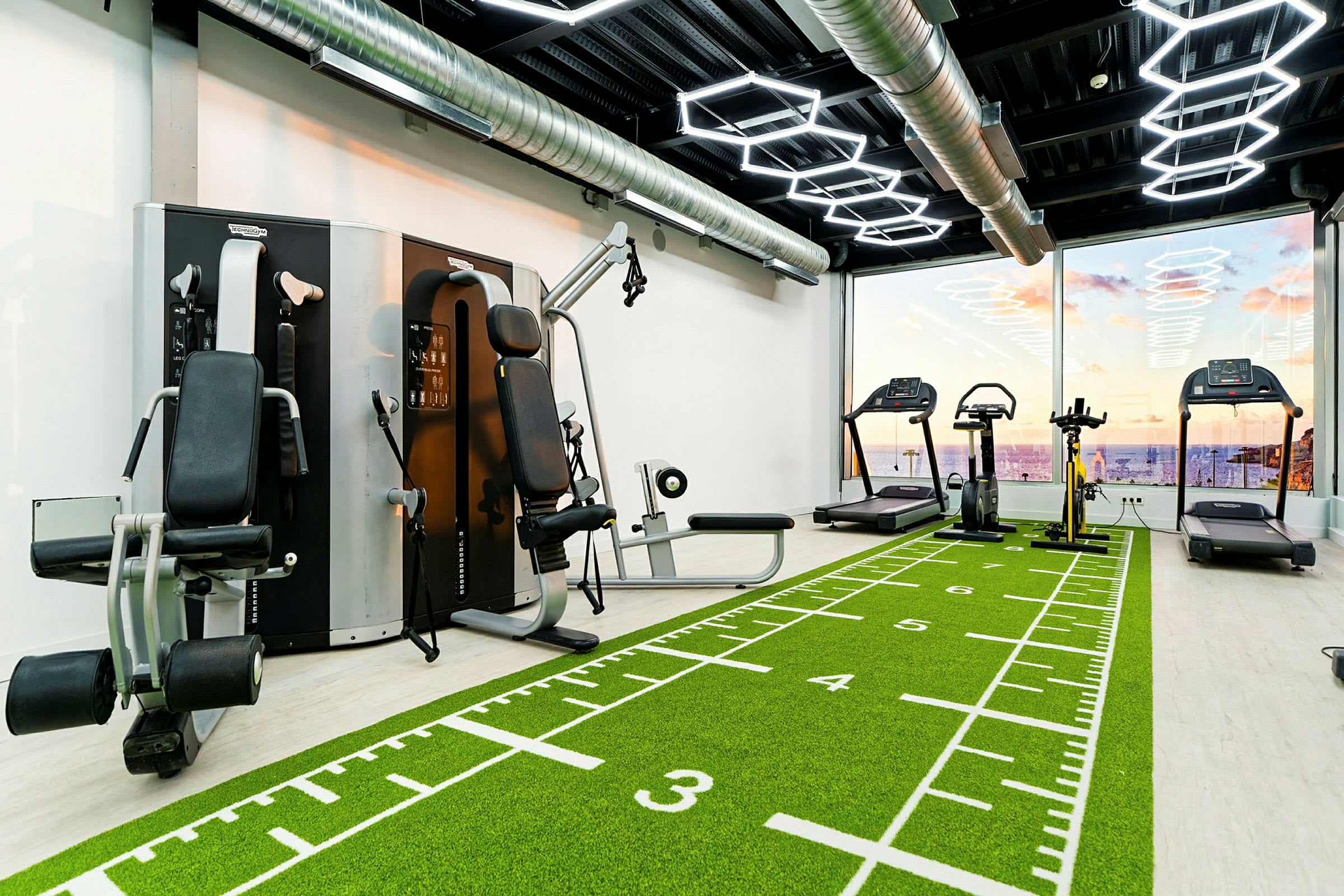 Ett gym med träningsmaskiner, löpband och grönt golv med markeringar. Stora fönster visar en utsikt över havet och en solnedgång. (AI-genererad text)