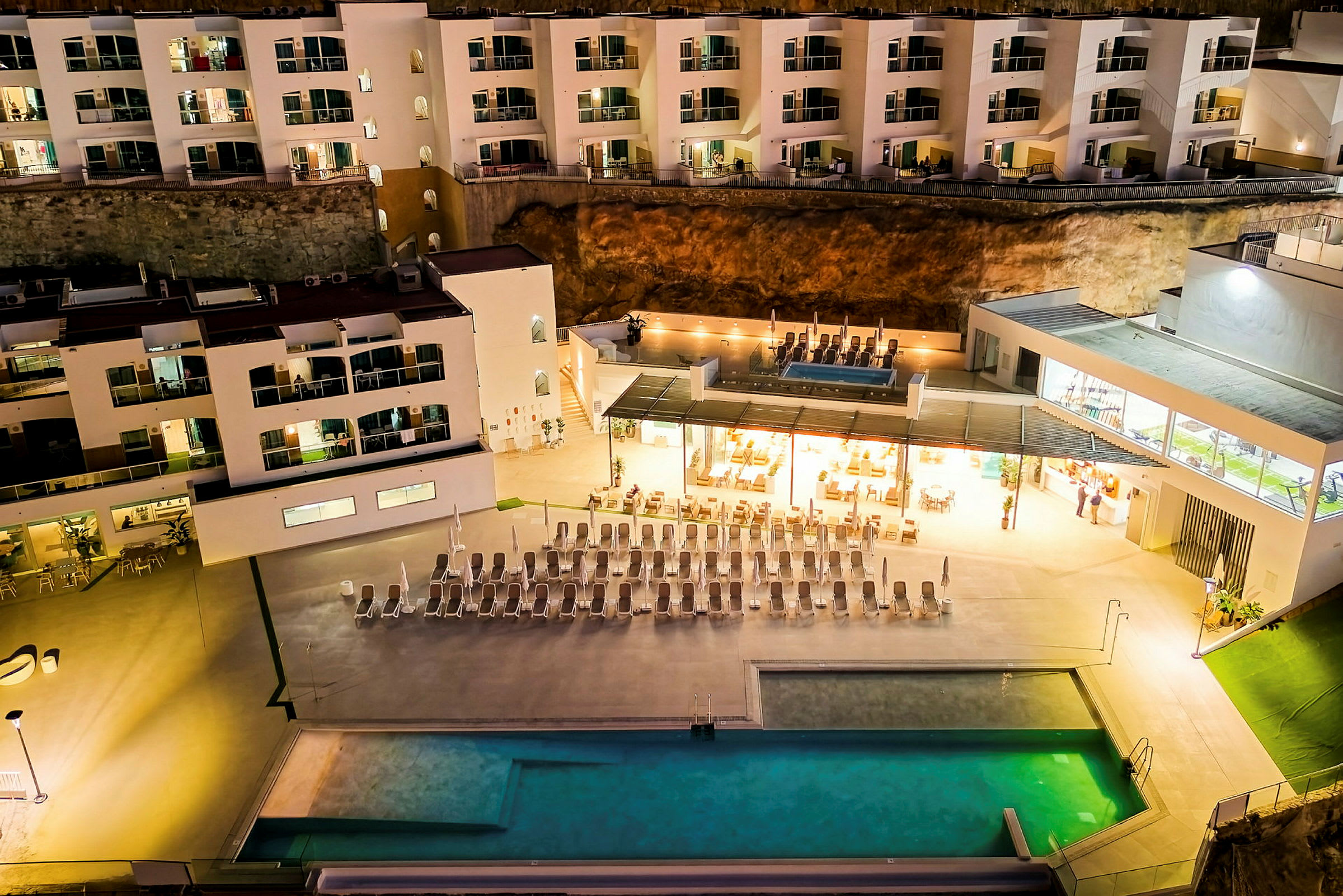 En byggnad på kvällen med integrerad pool och solstolar, samt ett upplyst restaurangområde. (AI-genererad text)