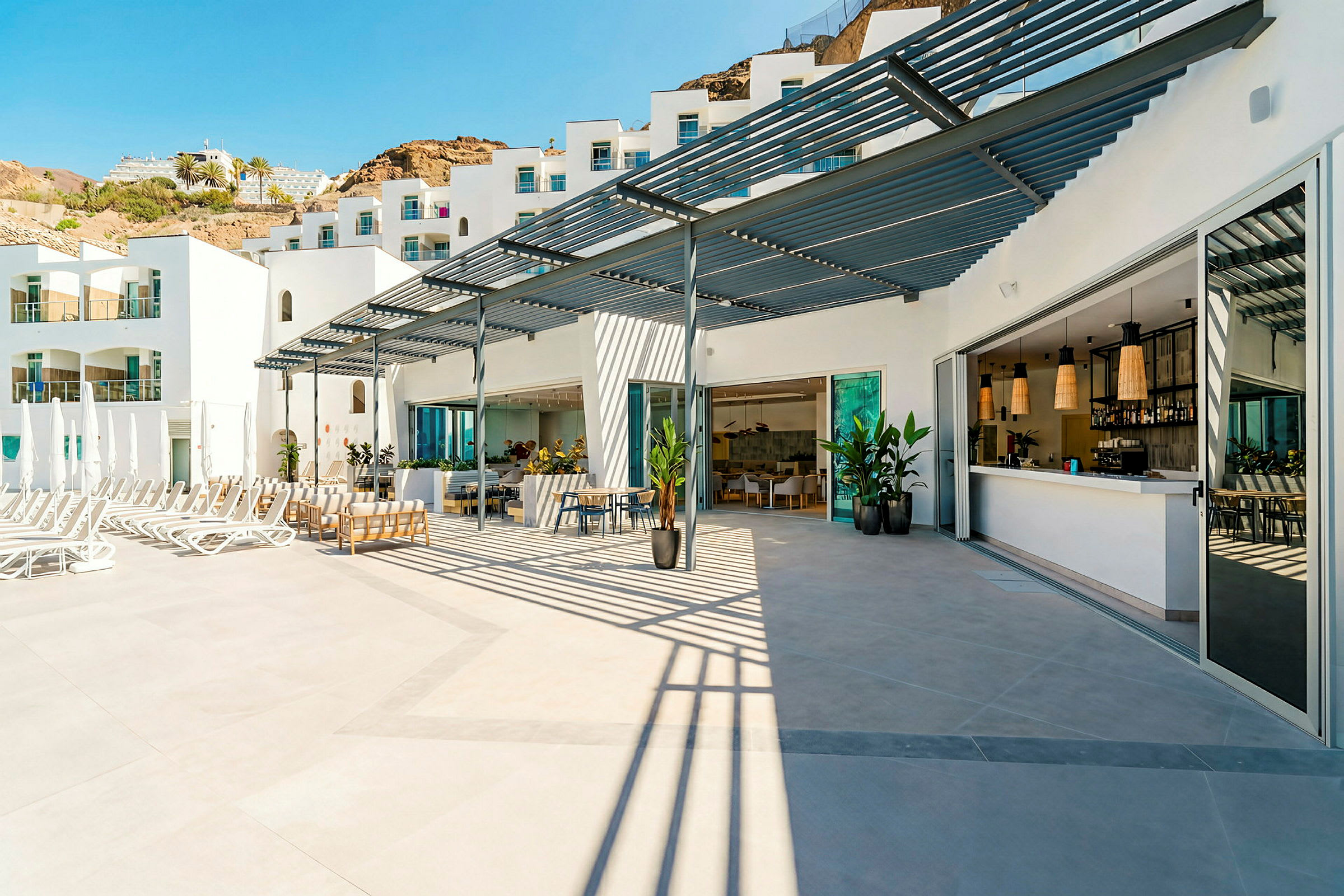 En utomhusterrass med solstolar, pergola och serveringsområde framför en vit byggnad. (AI-genererad text)