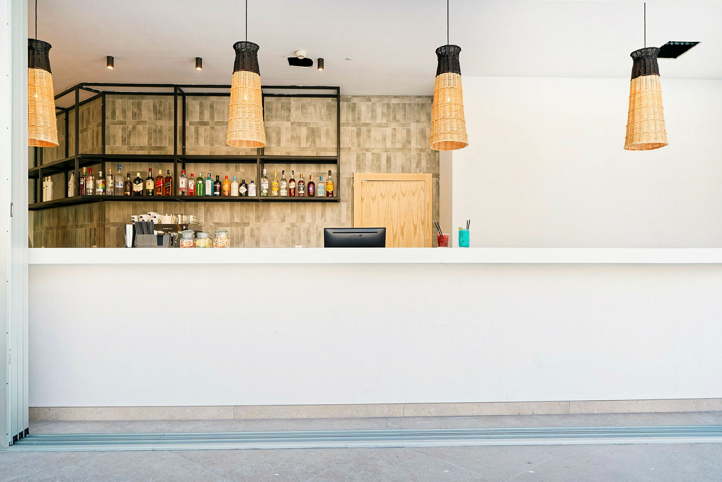 En modern bar med en vit disk, flaskor uppställda på hyllor i bakgrunden och tre hängande lampor. (AI-genererad text)