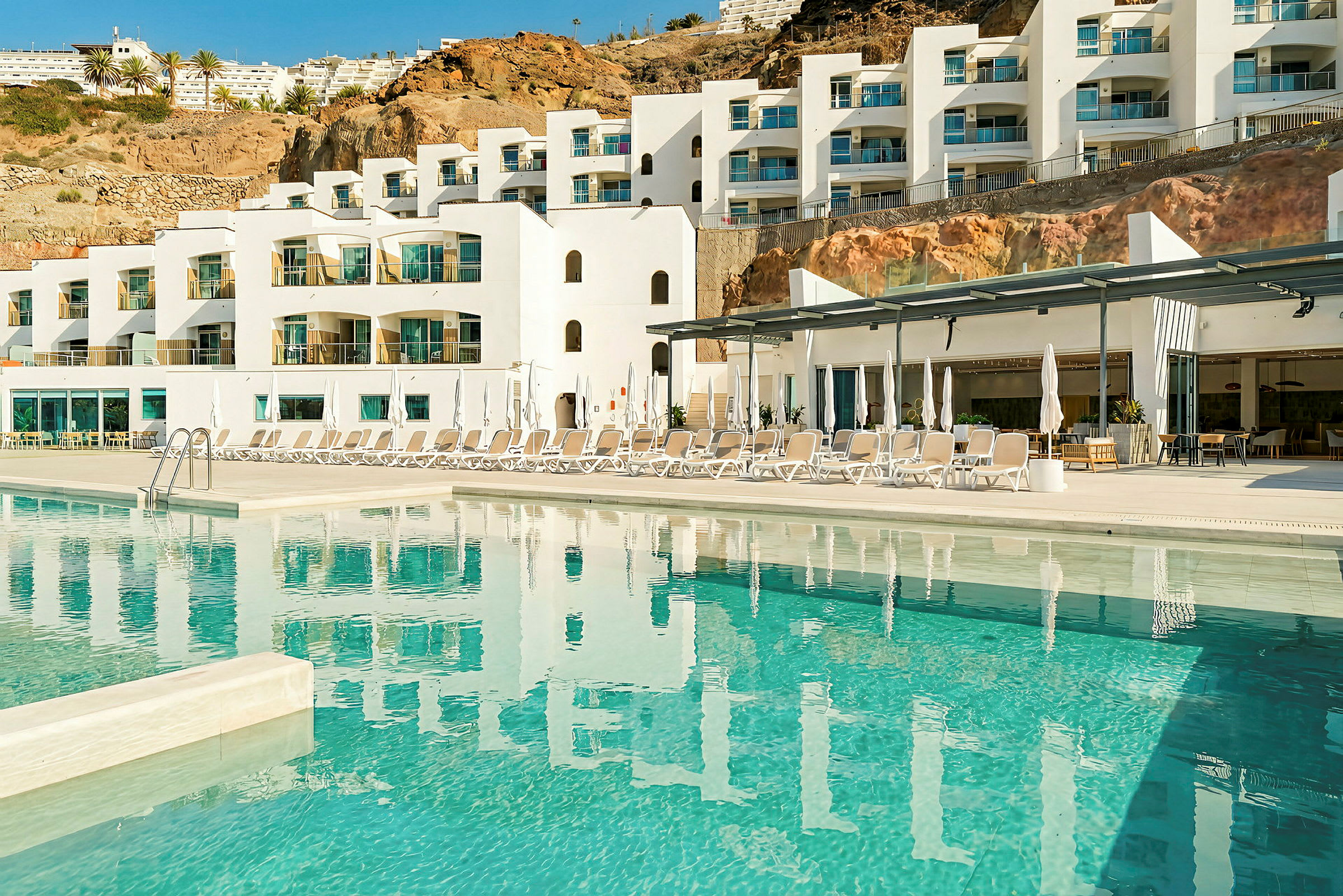 Ett modernt hotell med vit fasad och flera balkonger. Framför hotellet finns en pool och solstolar. (AI-genererad text)