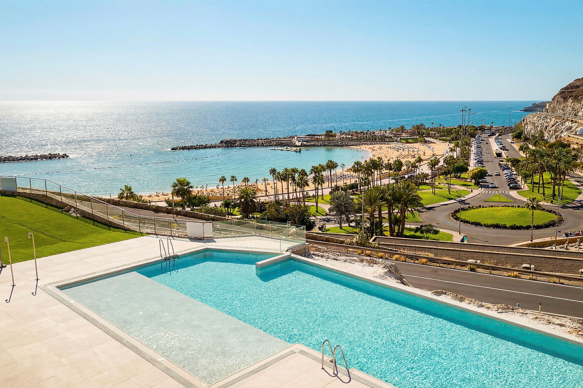 En swimmingpool med utsikt över en sandstrand, palmer och havet under en klarblå himmel. (AI-genererad text)