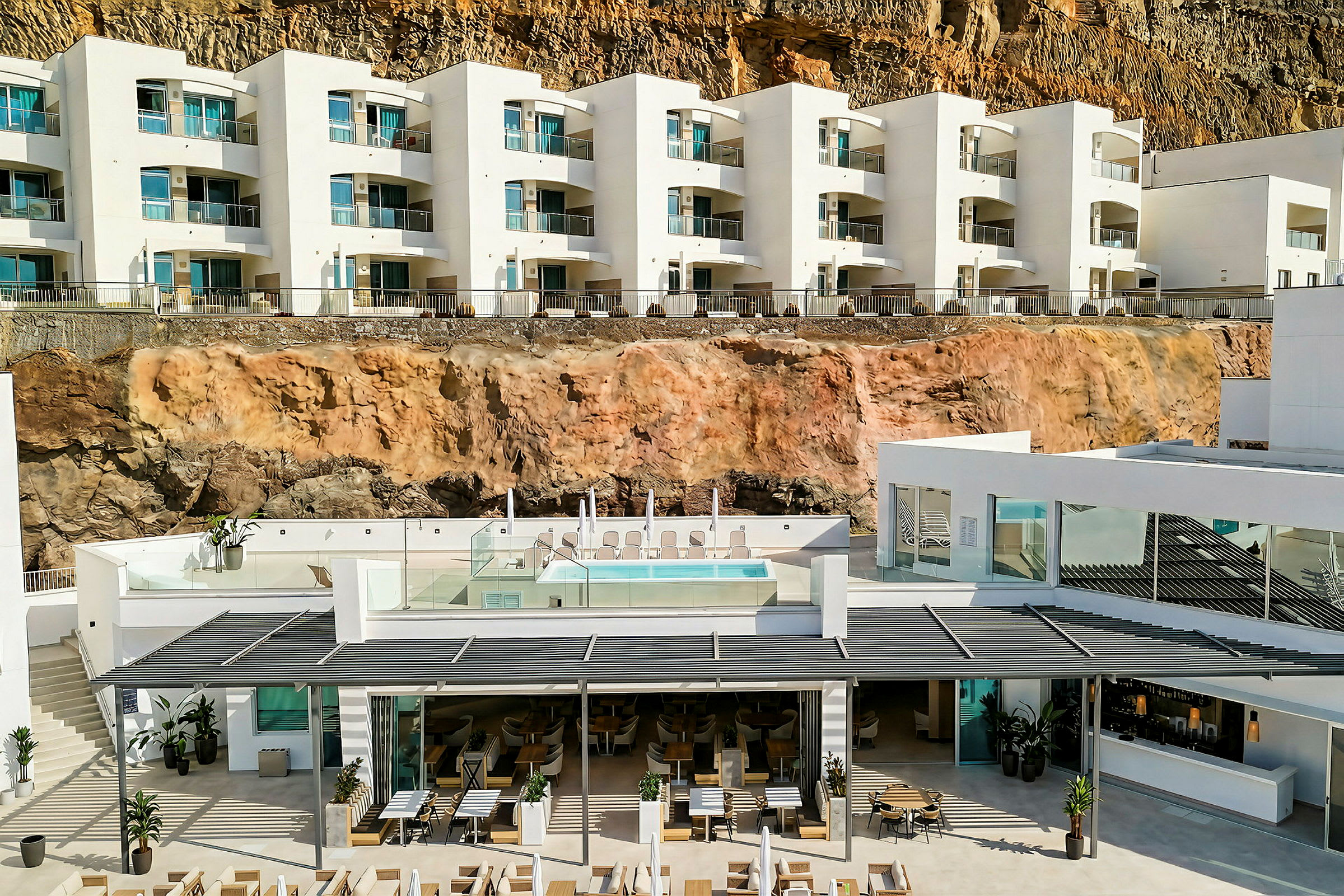 Ett modern hotell med vita fasader byggt vid en klippa. På det mellersta planet syns en pool och solstolar. (AI-genererad text)