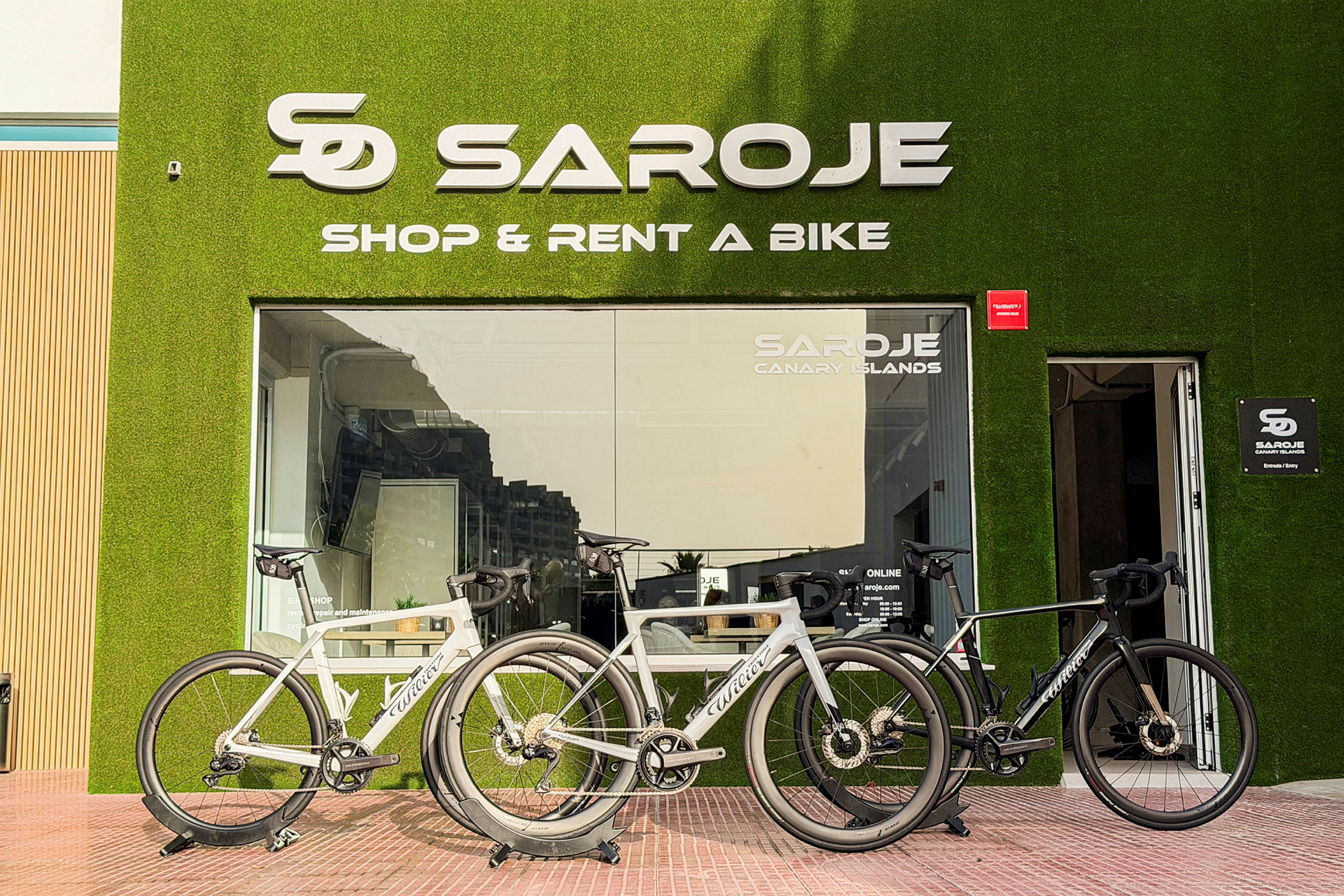 Tre moderna cyklar står parkerade framför en cykeluthyrningsbutik med grön fasad och glasfönster. (AI-genererad text)