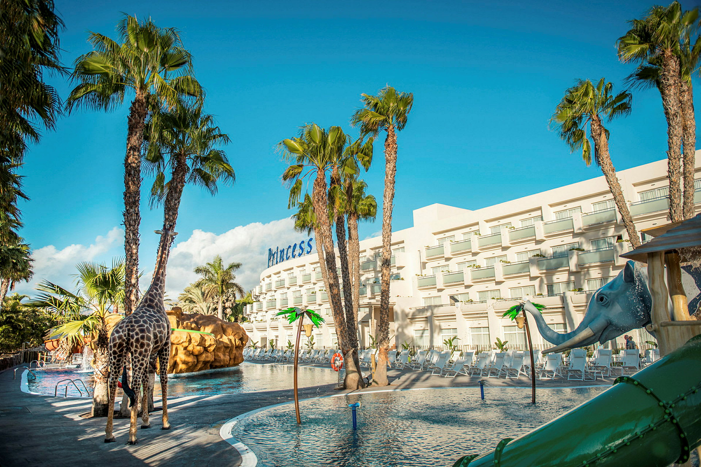 En pool med vattenrutschkana och leksaker i form av en giraff och en elefant, omgivna av palmer och en hotellbyggnad i bakgrunden. (AI-genererad text)