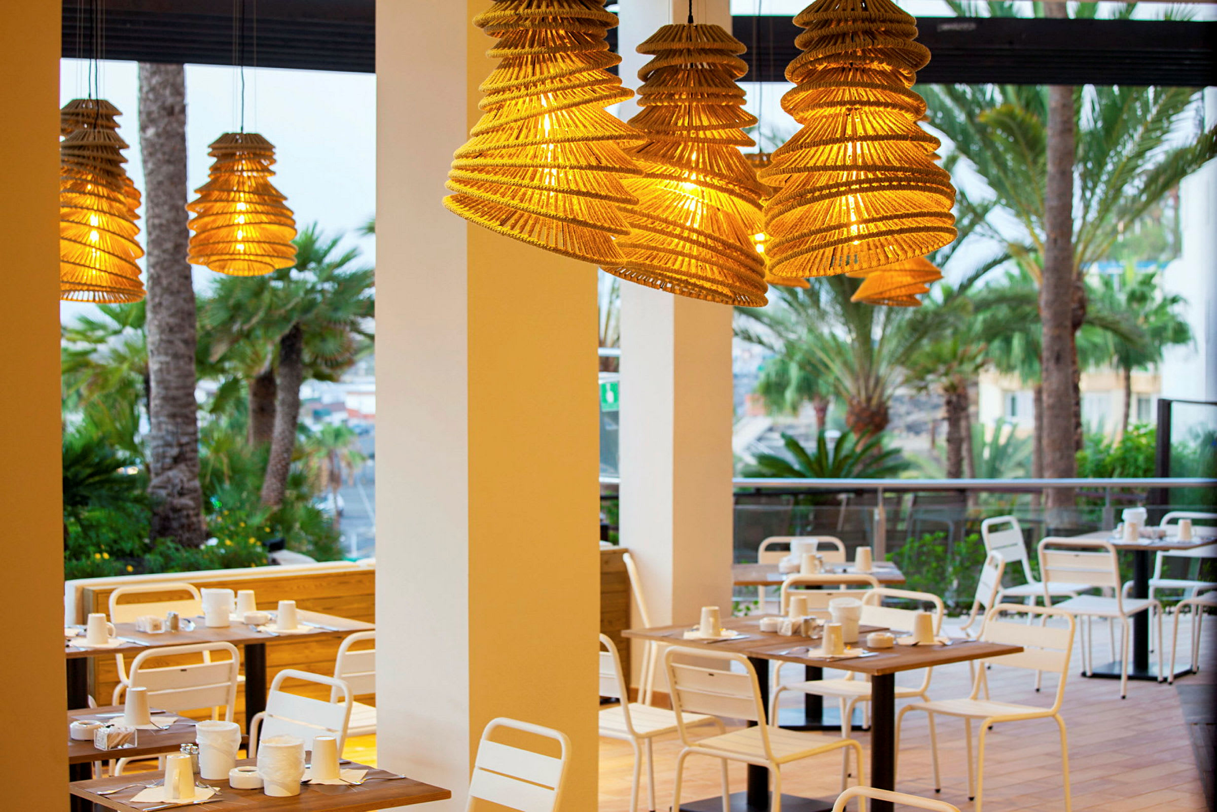 Utemöbler vid restaurang med hängande lampskärmar av trä och palmträd i bakgrunden. (AI-genererad text)