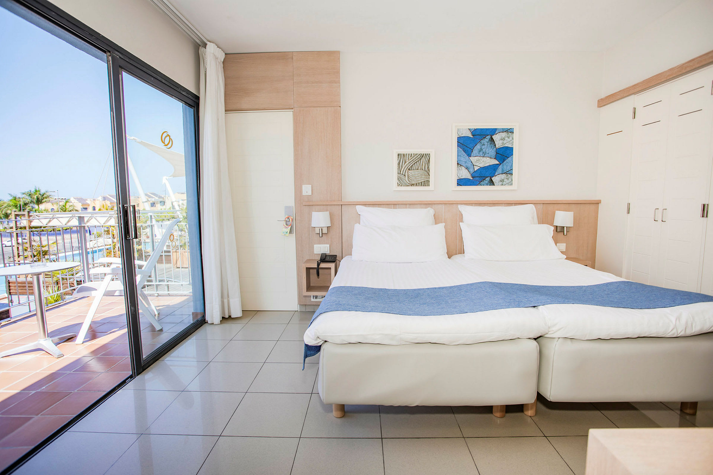 Ett ljust hotellrum med två enkelsängar som är sammanfogade. Det finns ett glasparti som leder ut till en balkong med utsikt över ett poolområde. (AI-genererad text)