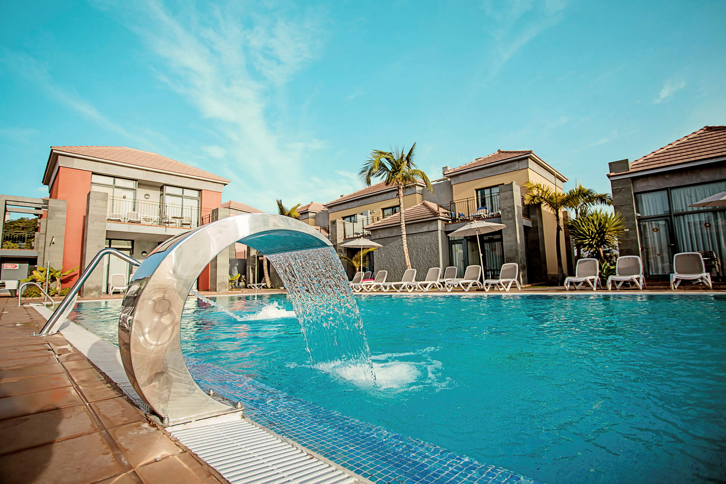 En swimmingpool med en modern vattenfontän omgiven av vilstolar och semesterhus under klarblå himmel. (AI-genererad text)