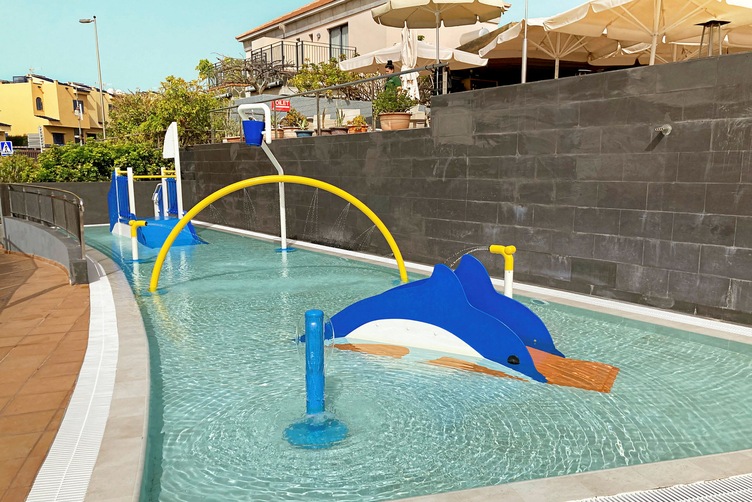 Barnpool med lekredskap såsom en blå delfin och en gul båge som sprutar vatten. (AI-genererad text)