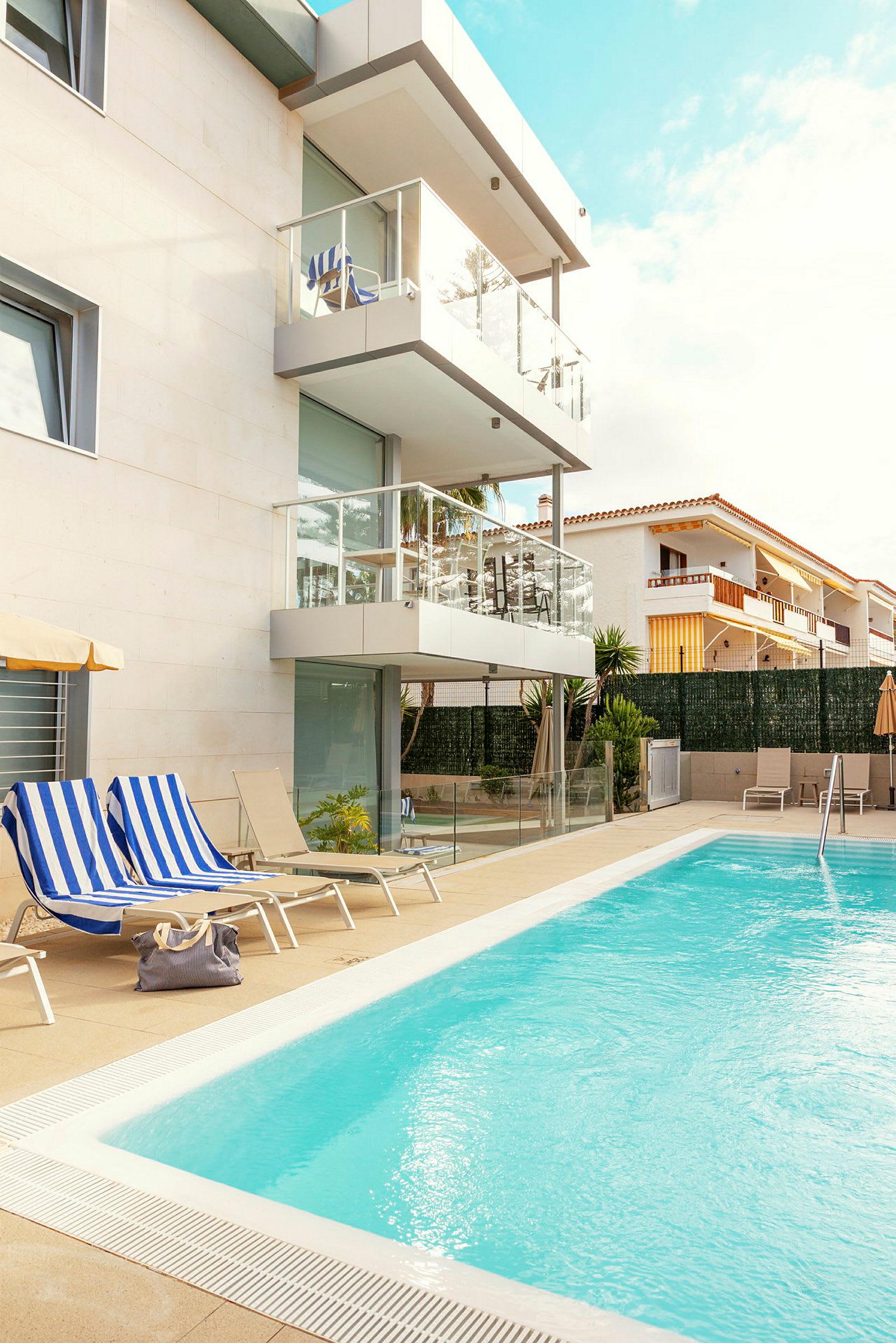 Bild på en pool intill en modern byggnad med balkonger, solstolar och en blå himmel. (AI-genererad text)