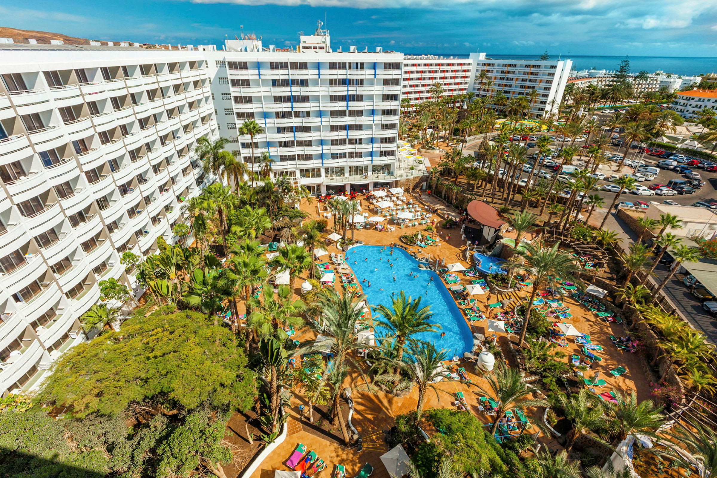 Flygfoto av en semesterresort med pool, omgiven av solstolar, palmer och flerbostadshus. (AI-genererad text)
