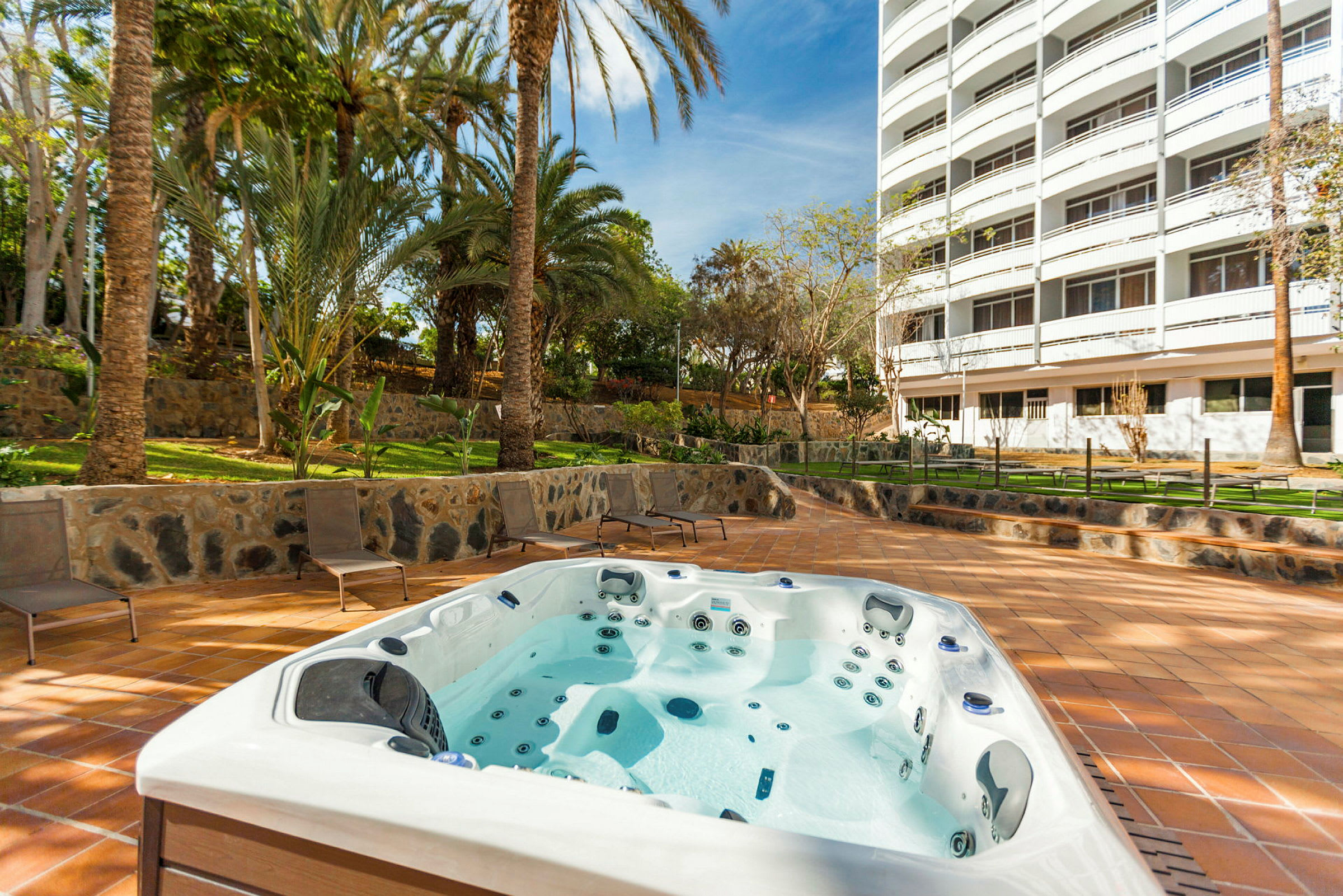 Bild av en utomhus jacuzzi belägen nära en byggnad med flera våningar och omgiven av grönska och palmer. (AI-genererad text)