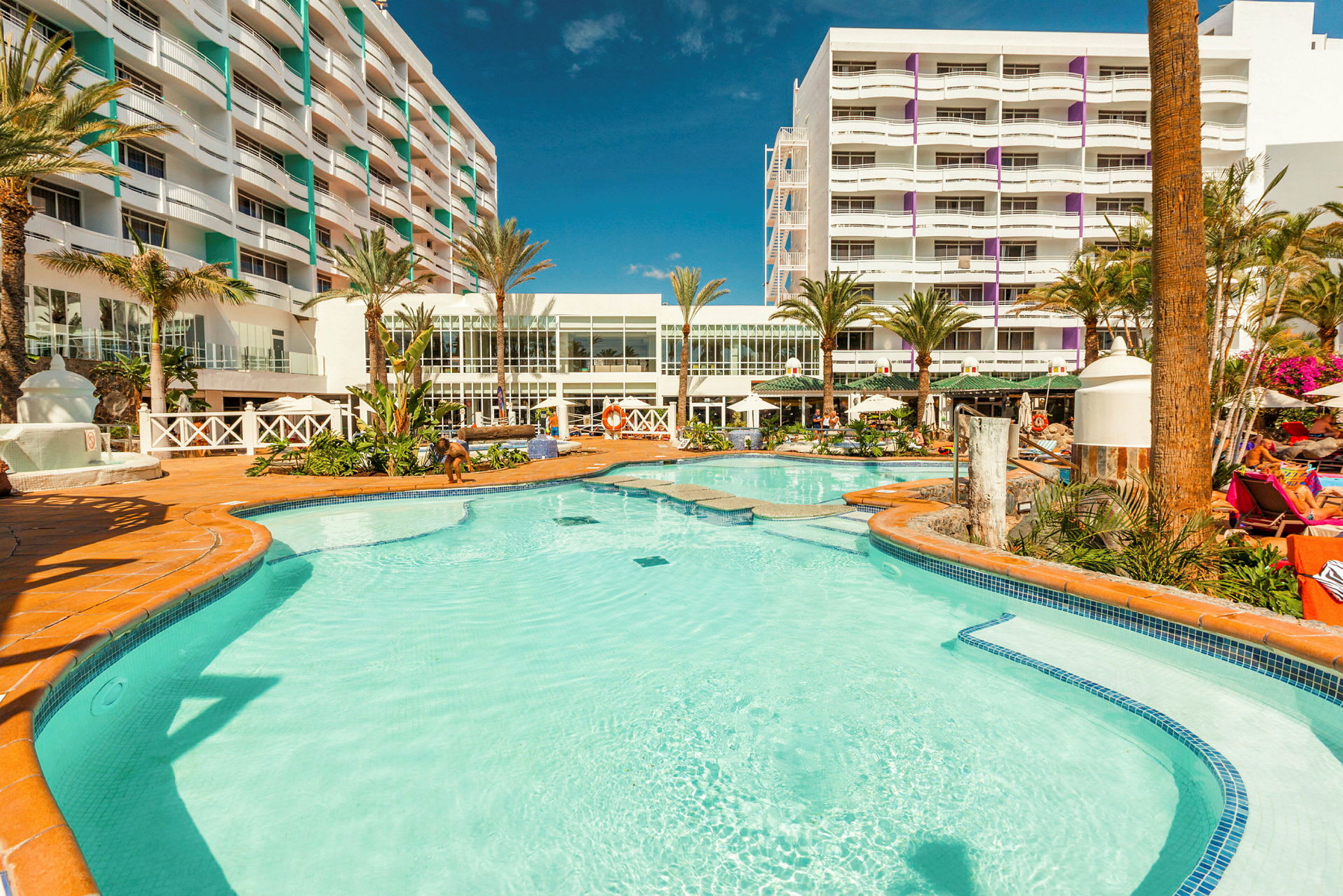 Utomhuspool med omgivande solstolar och hotelbyggnad i bakgrunden. (AI-genererad text)