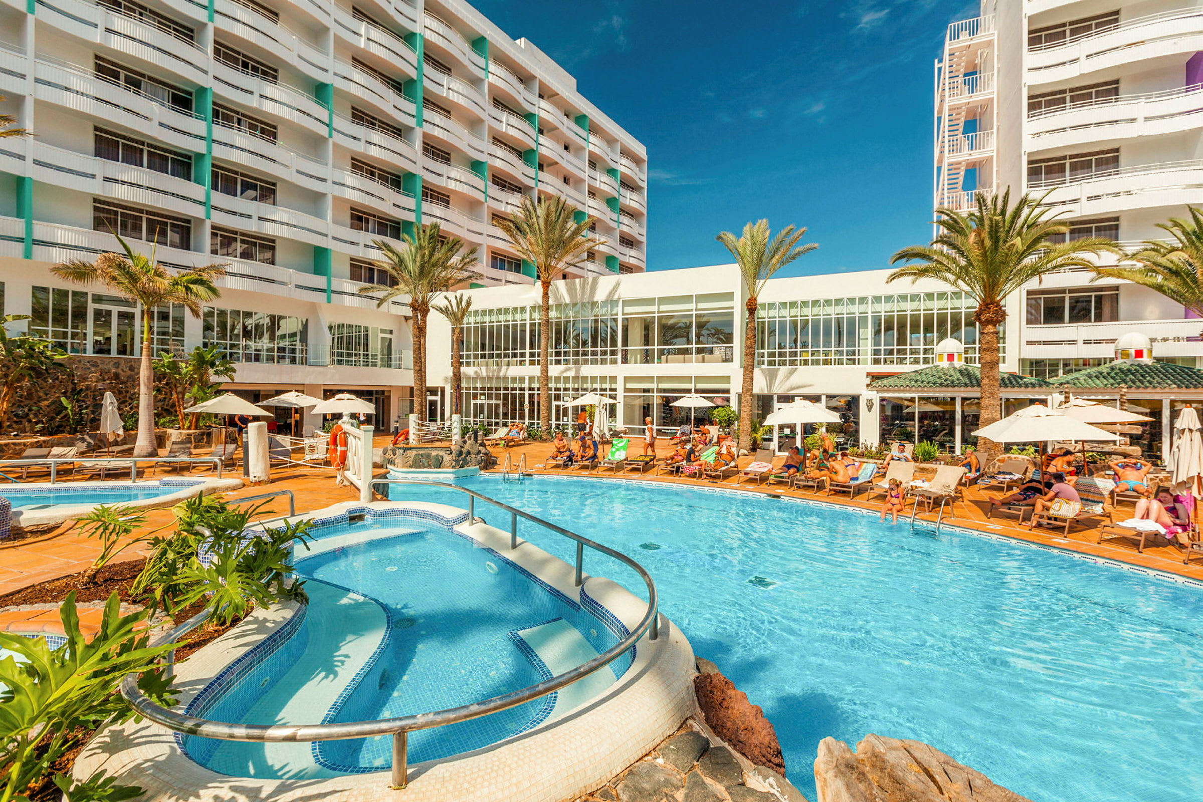Hotellområde med pool, solstolar och byggnad i bakgrunden. (AI-genererad text)