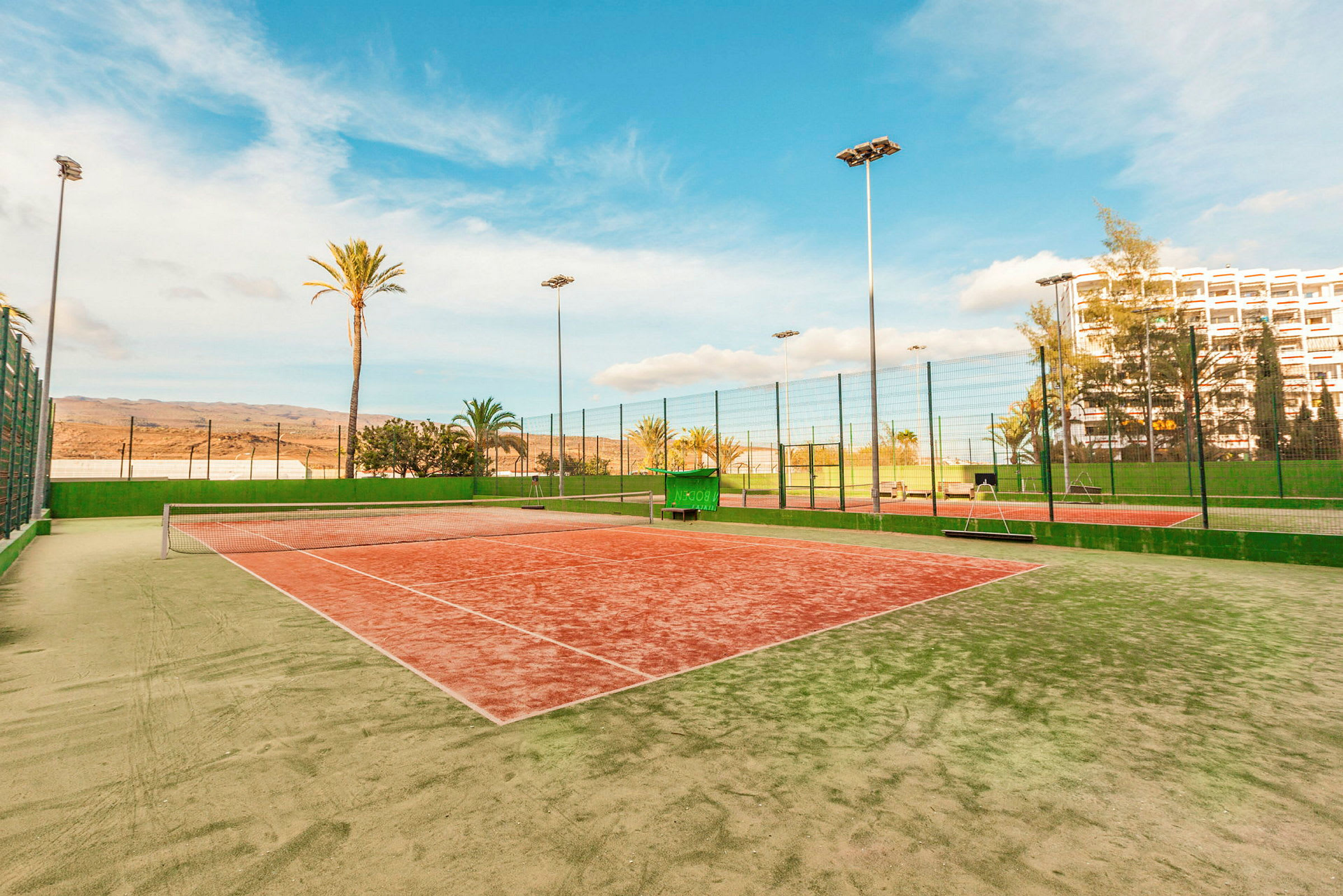 En tennisbana med grön och röd yta, omgiven av höga nät och palmer. (AI-genererad text)