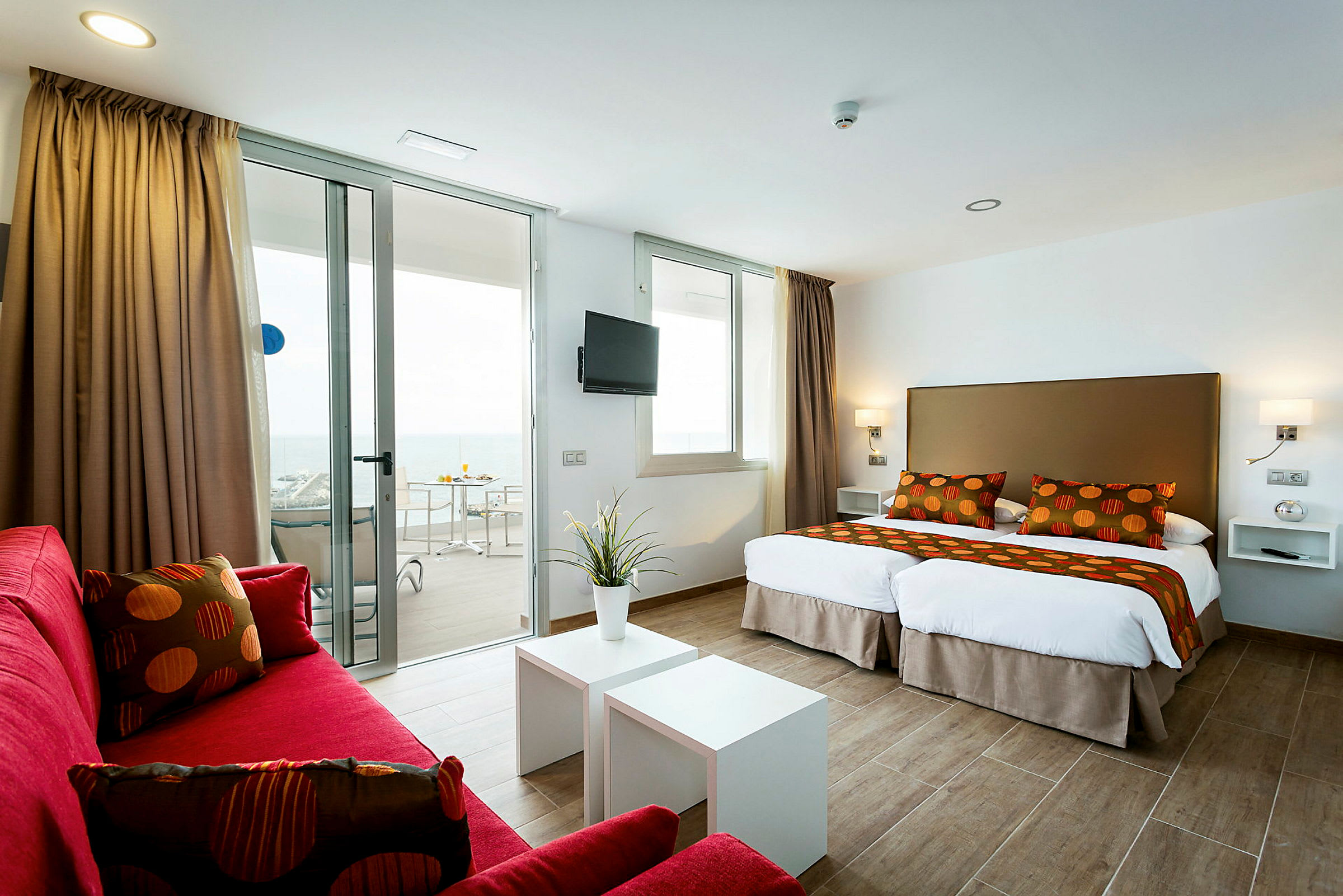 Ett modernt hotellrum med två enkelsängar, röda soffmöbler och en balkong med havsutsikt. (AI-genererad text)