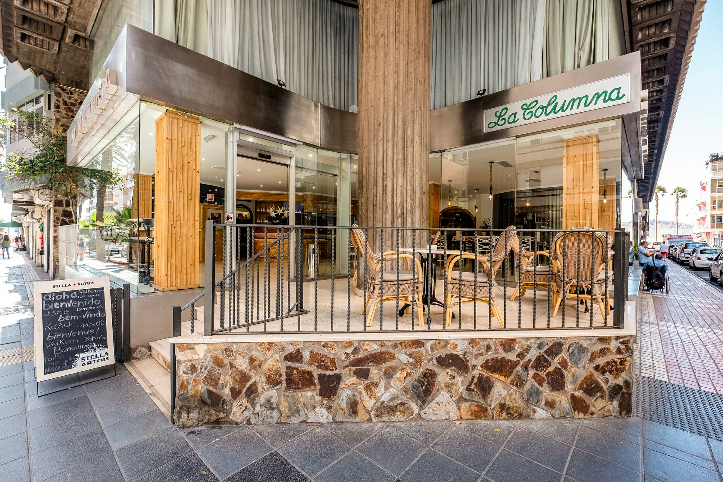 En restaurangs entré med uteservering där det står bord och stolar. Restaurangen heter "La Columna" och ligger vid en trottoar. (AI-genererad text)